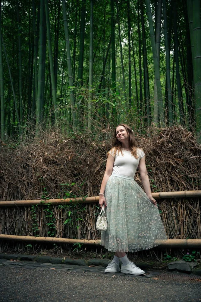A young woman standing in front of a bamboo forest, wearing a white top, a light green floral skirt, and white sneakers, holding a small white bag, with a relaxed smile.