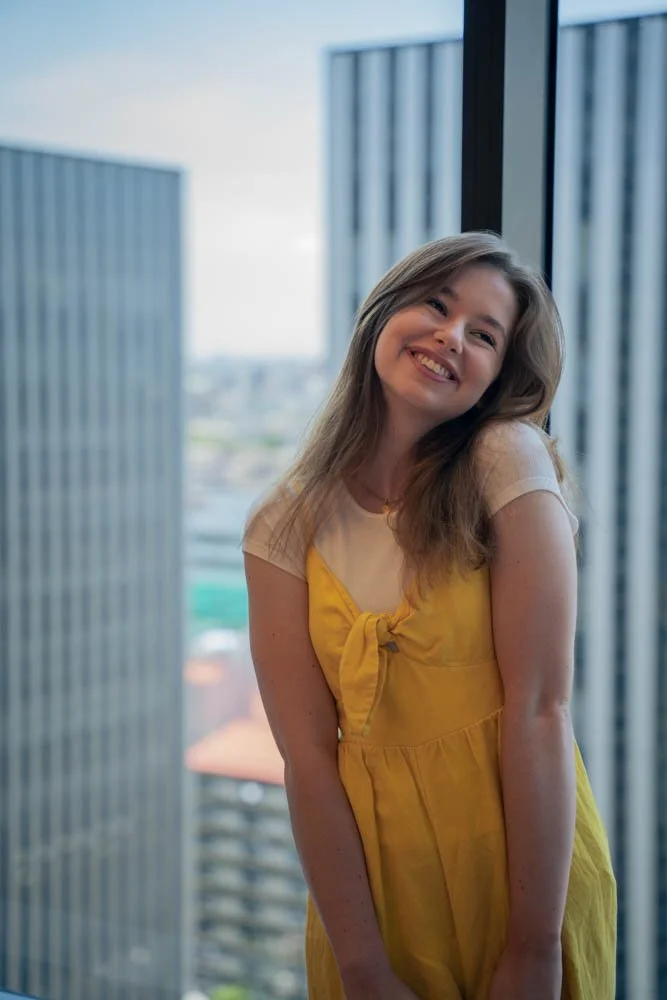 A young woman with light brown hair smiling and posing in front of a large window in a high-rise building, wearing a yellow dress with a front knot detail.