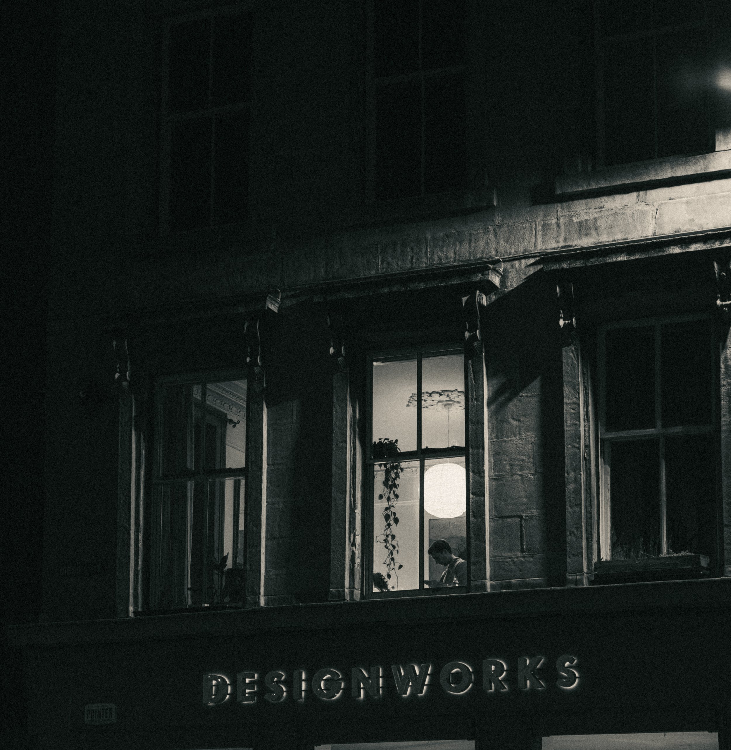 Nighttime view of a building with a large illuminated sign reading 'DESIGNWORKS' and part of another sign at the bottom. The building has a dark brick exterior with windows and some plants visible inside.