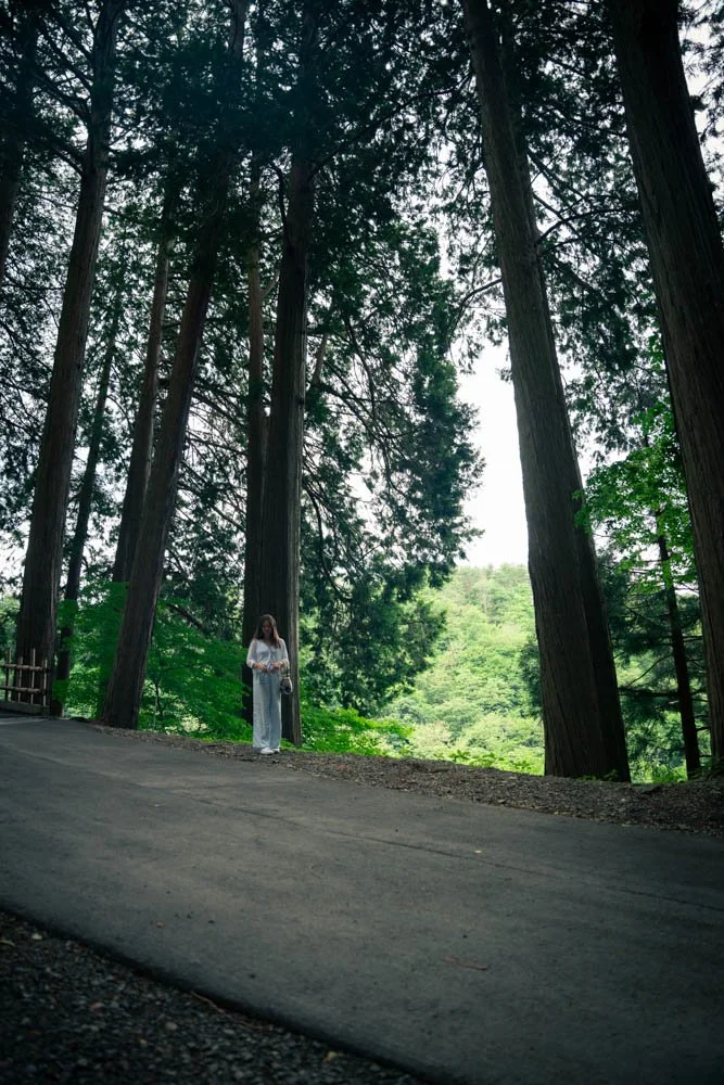 A woman in a white shirt and light-colored pants stands on a paved path in a forest with tall trees and green foliage.