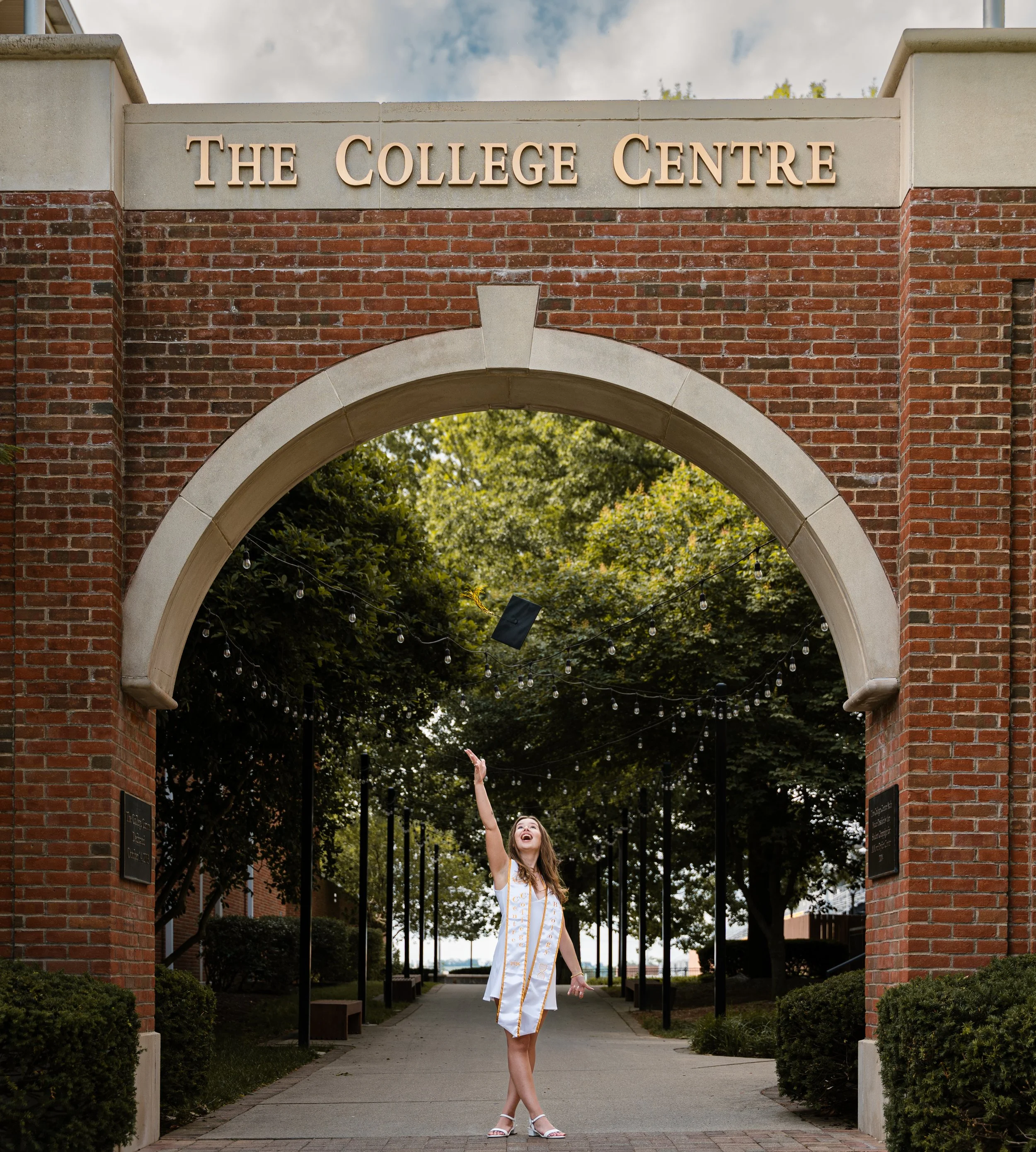 A young woman in a white and gold graduation gown is standing with one arm raised beneath an archway that reads 'The College Centre.' She is smiling and looking upward, with trees and clouds in the background.
