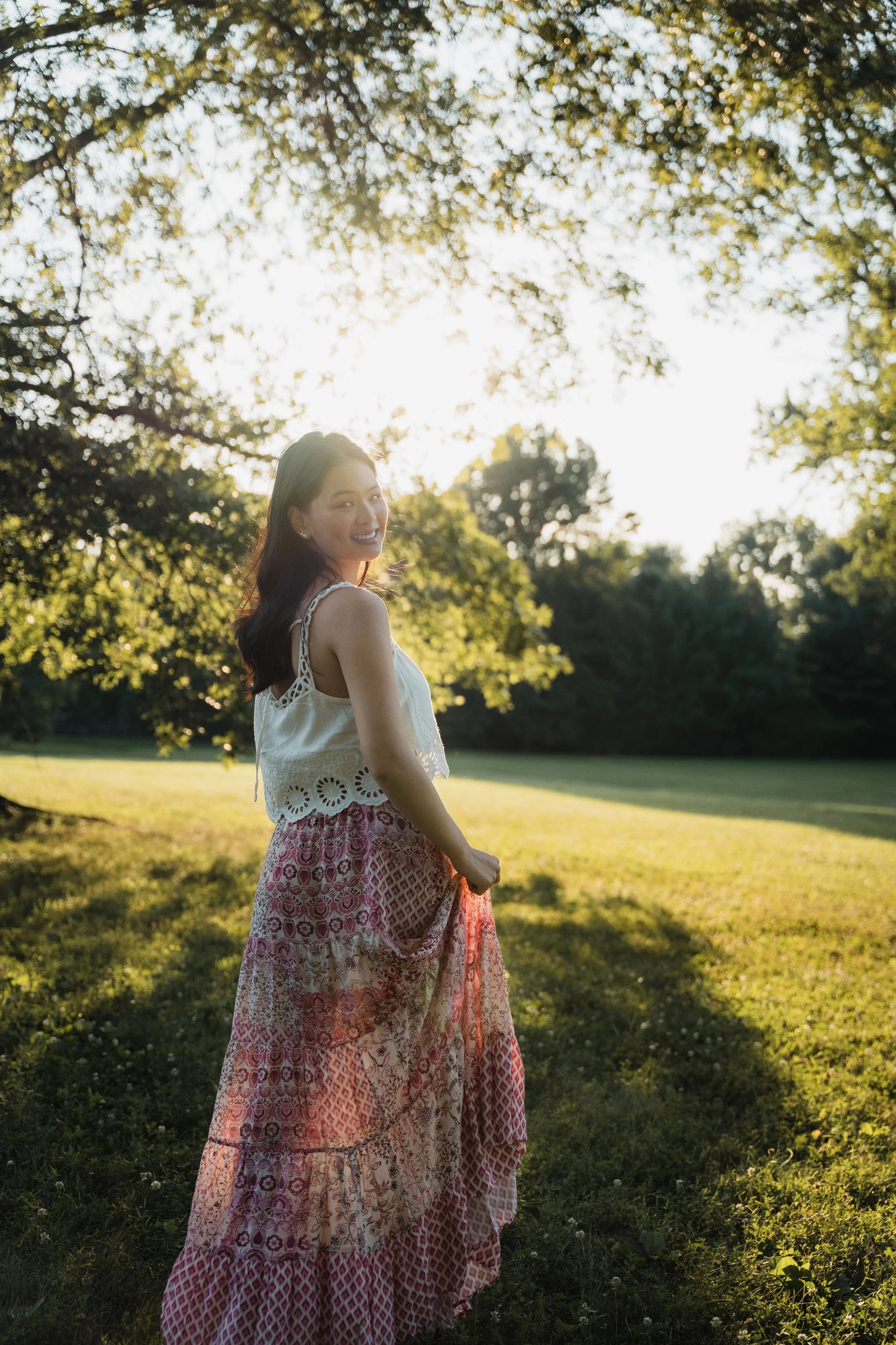 A woman standing in a grassy park, smiling, with trees and sunlight in the background.