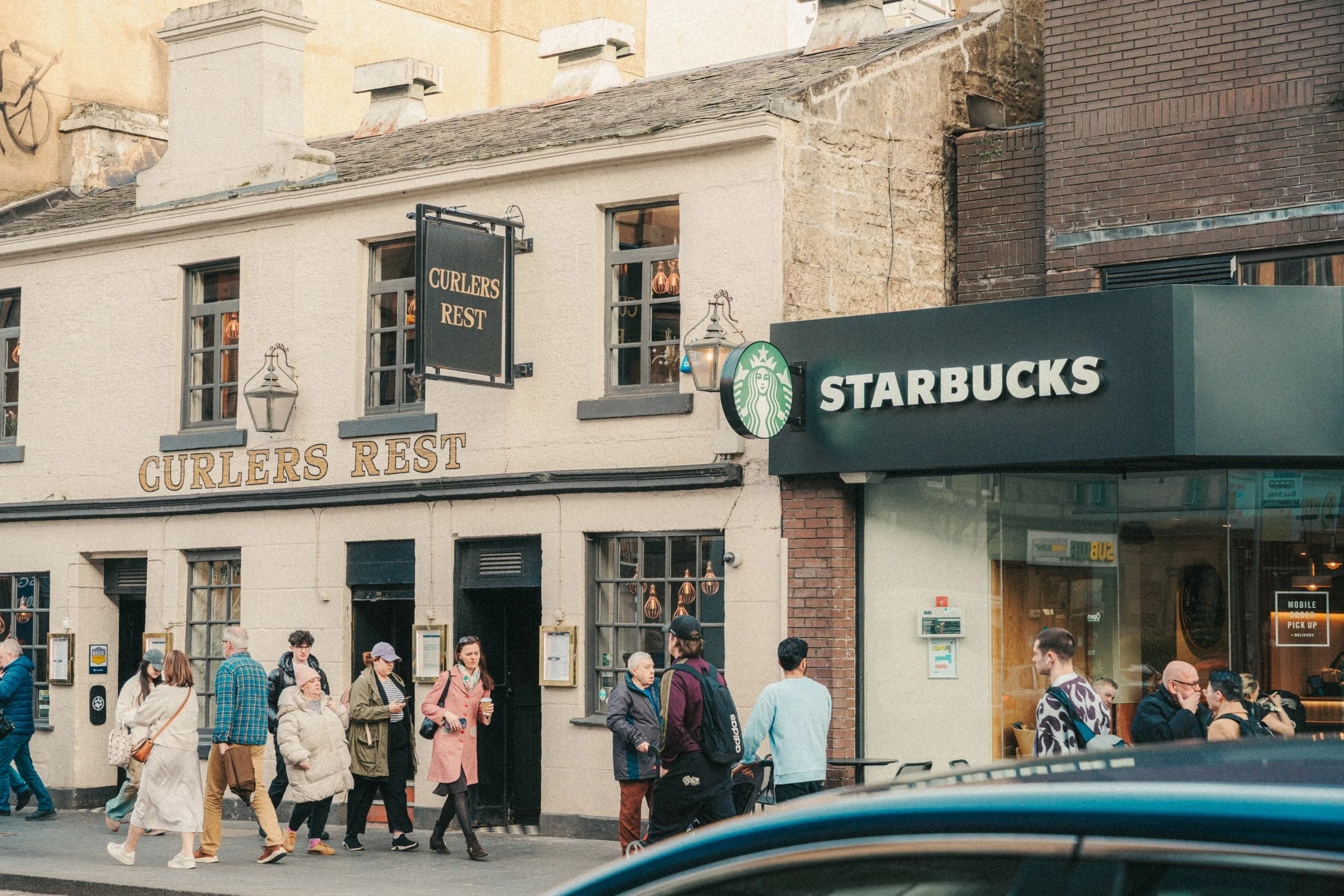 People walking on the sidewalk in front of a building with signs for "Curlers Rest" and Starbucks, with a crowd gathered and engaged in various activities.