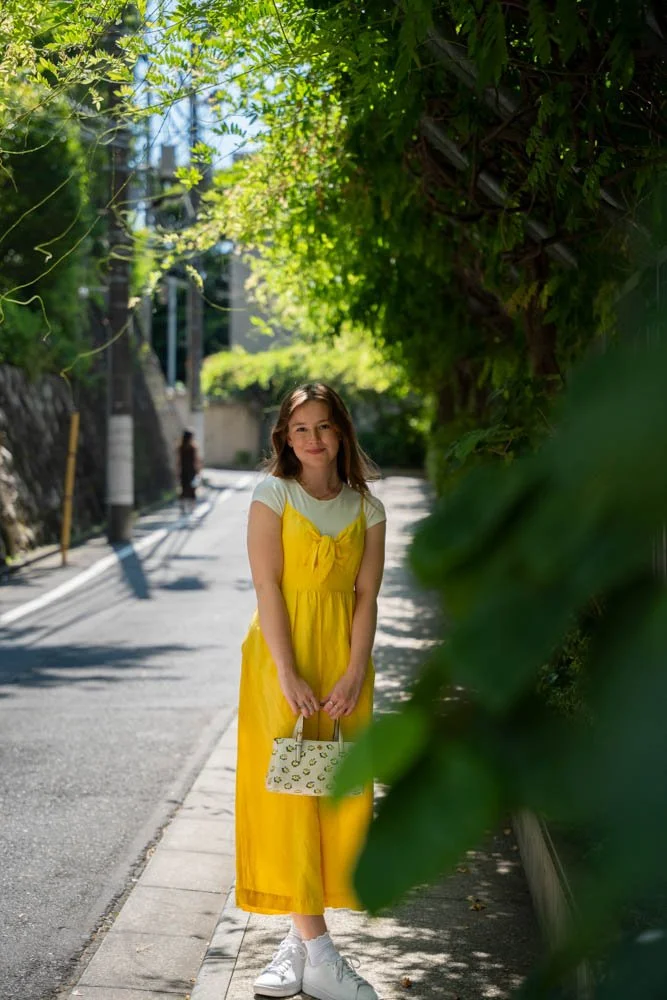 Young girl standing on sidewalk, wearing a yellow dress and white sneakers, holding a small patterned bag, smiling, with greenery and trees in the background on a sunny day.
