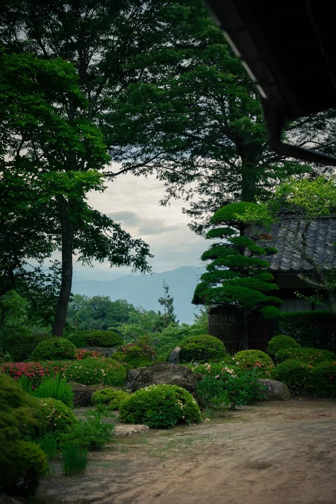 A traditional Japanese garden with lush green trees, neatly trimmed bushes, rocks, and a gravel pathway. A section of a wooden building with a tiled roof is visible on the right side, and in the background, misty mountains can be seen under an overca