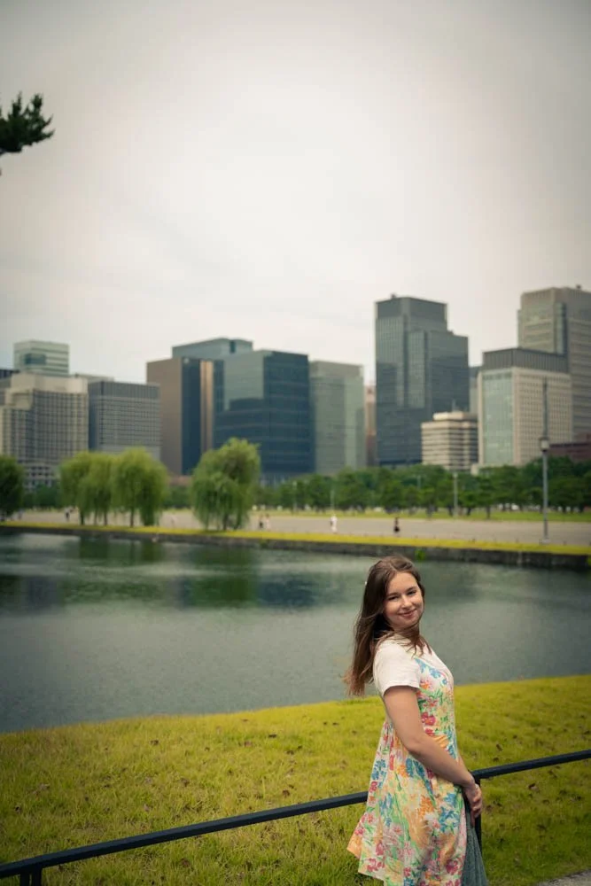 A young girl standing near a body of water in a park, with a city skyline of tall office buildings in the background.