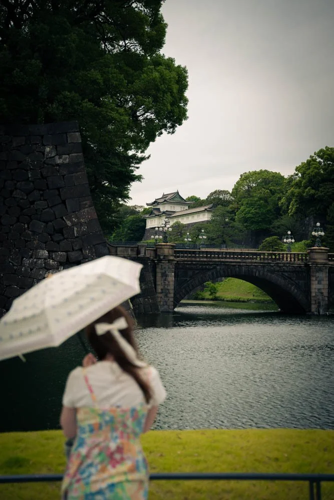 A young girl holding a white umbrella and standing near a river, with a bridge and traditional Japanese castle in the background under an overcast sky.