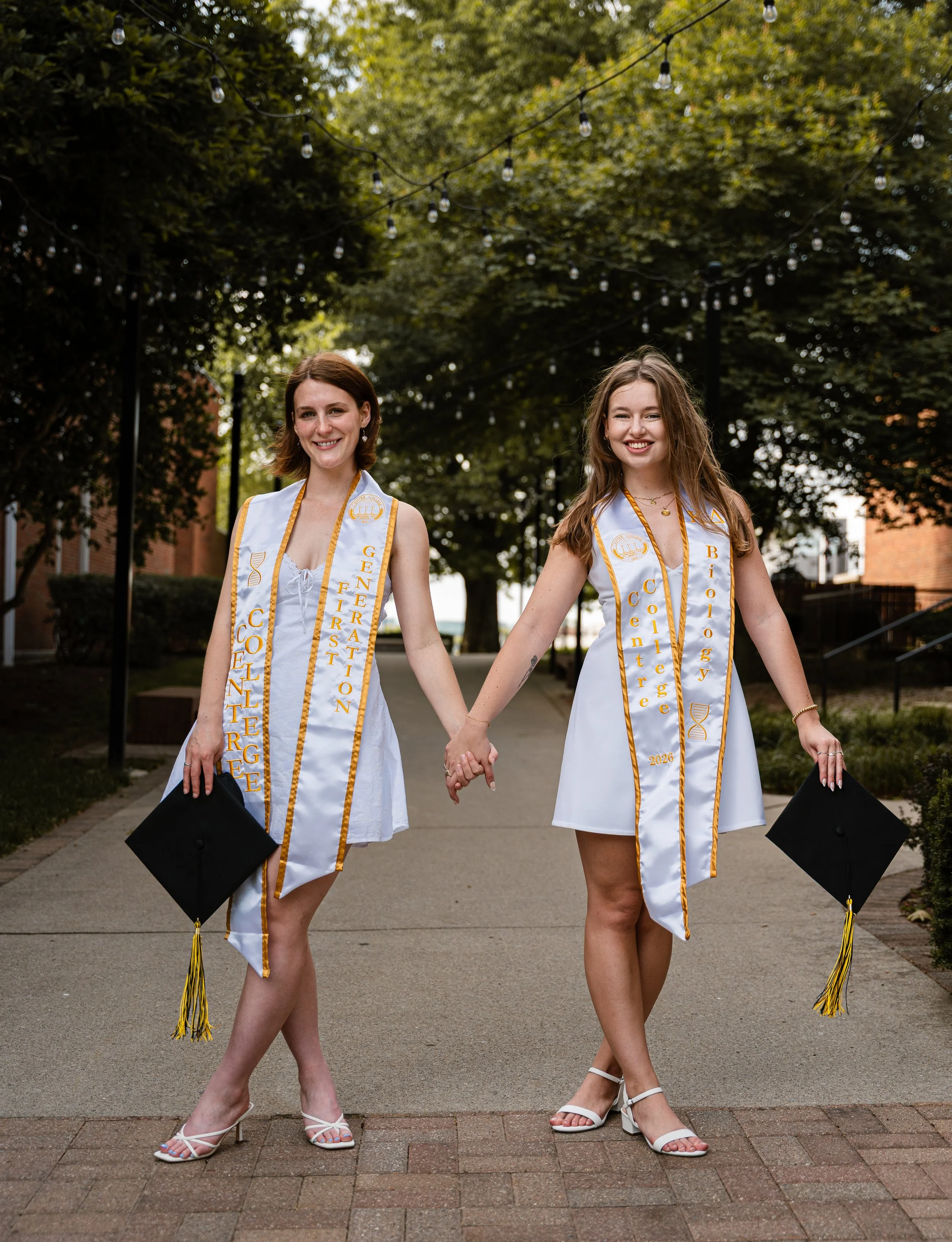 Two young women in white graduation dresses and heels, holding hands and carrying diplomas, standing outdoors beneath string lights and trees, smiling at the camera.