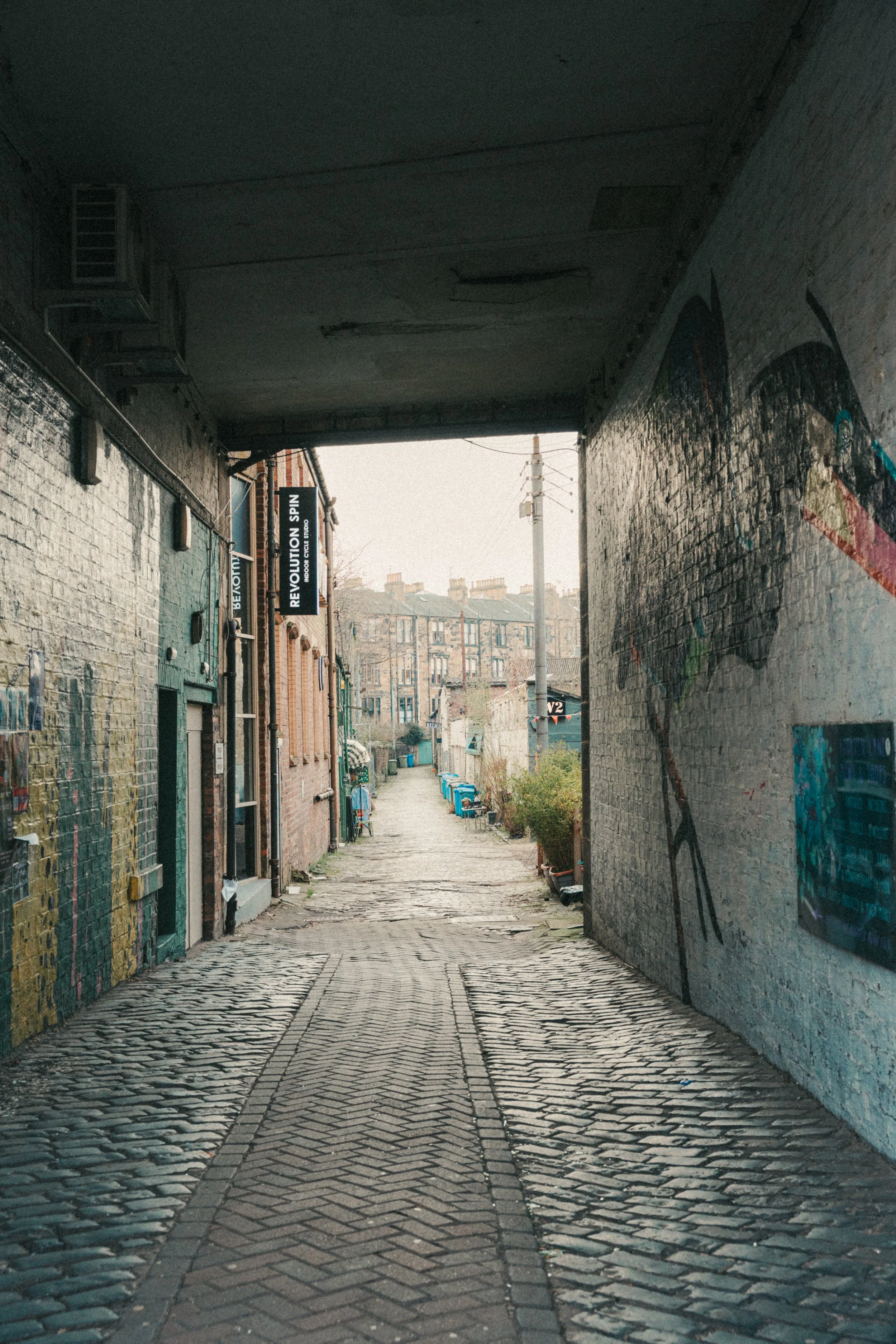 A narrow cobblestone alleyway between brick buildings, featuring street art on the walls and a line of blue recycling bins at the end.