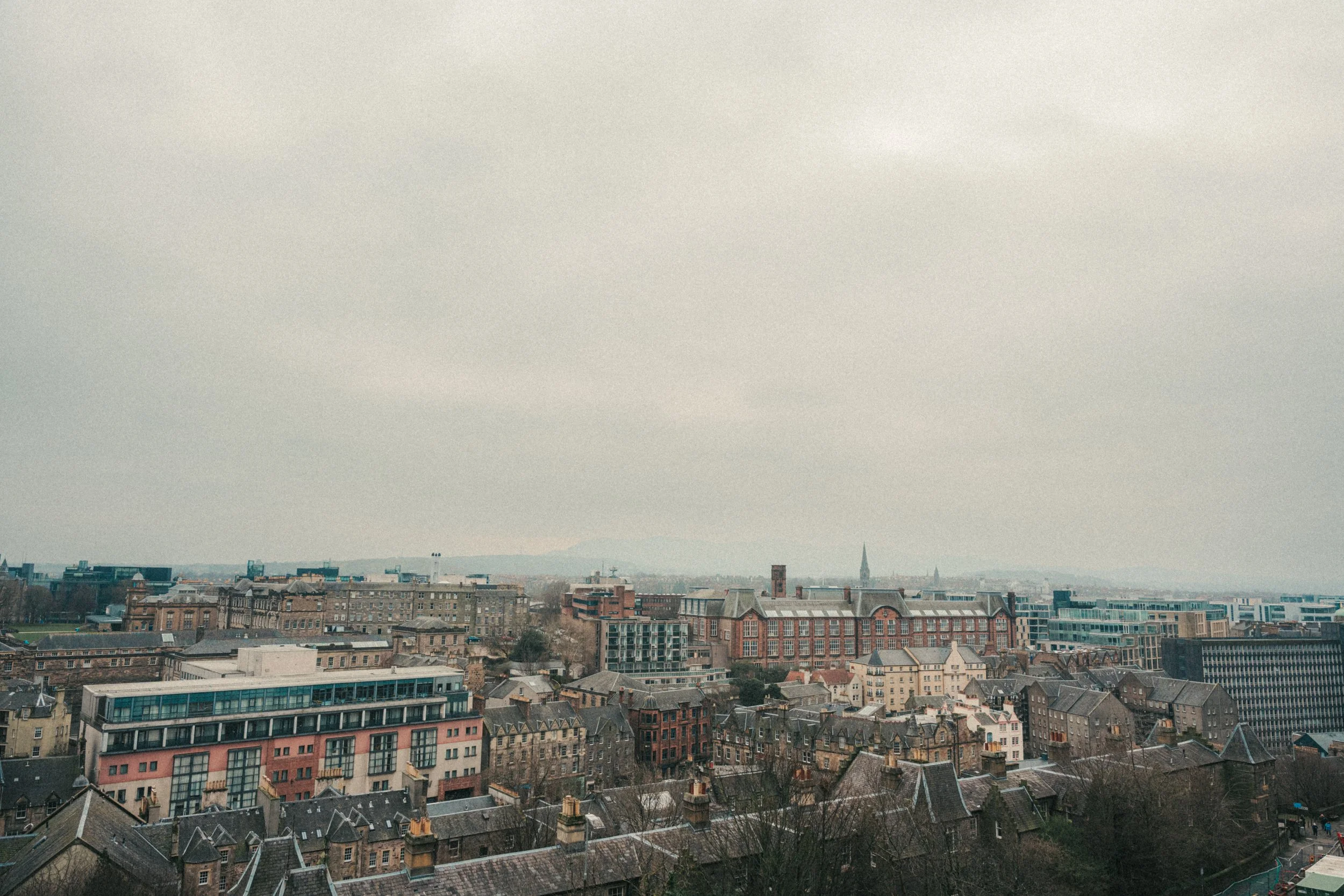 Cityscape view showing rooftops of various buildings and houses, with a few taller structures and a church steeple in the distance under an overcast cloudy sky.