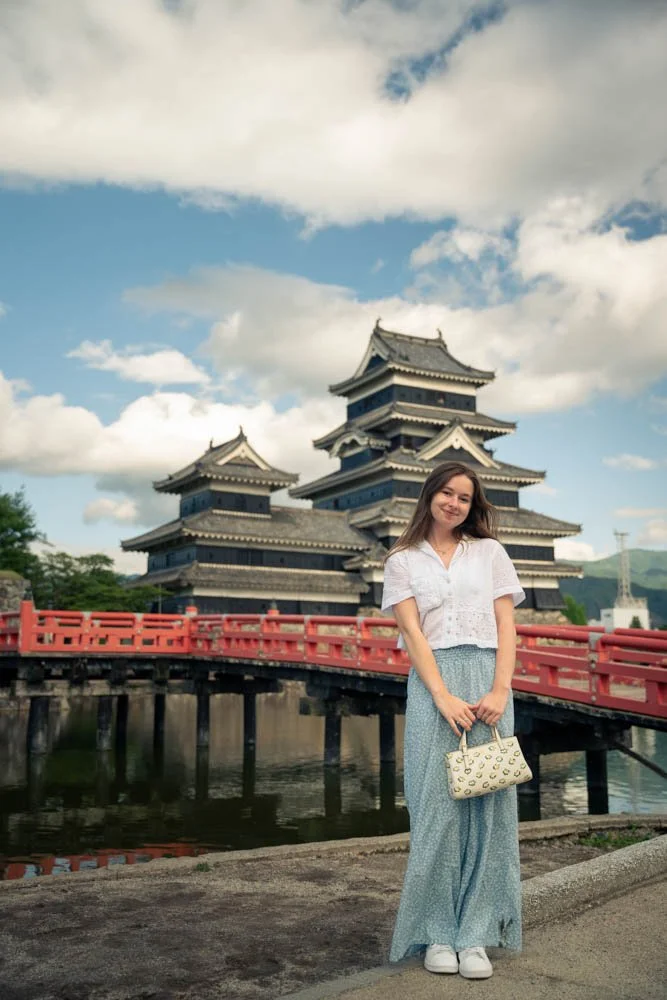 A young woman standing on a bridge in front of a traditional Japanese-style pagoda-style building with multiple tiers, surrounded by water and cloudy sky.