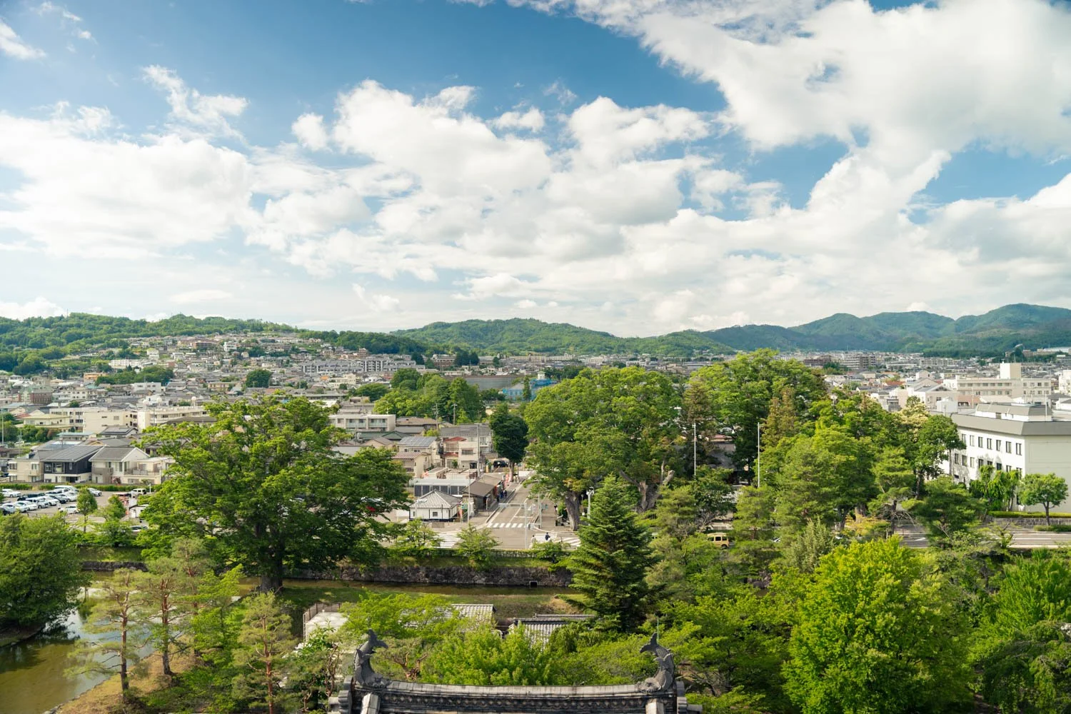 A panoramic view of a city with numerous trees, buildings, and hills in the background under a partly cloudy sky.