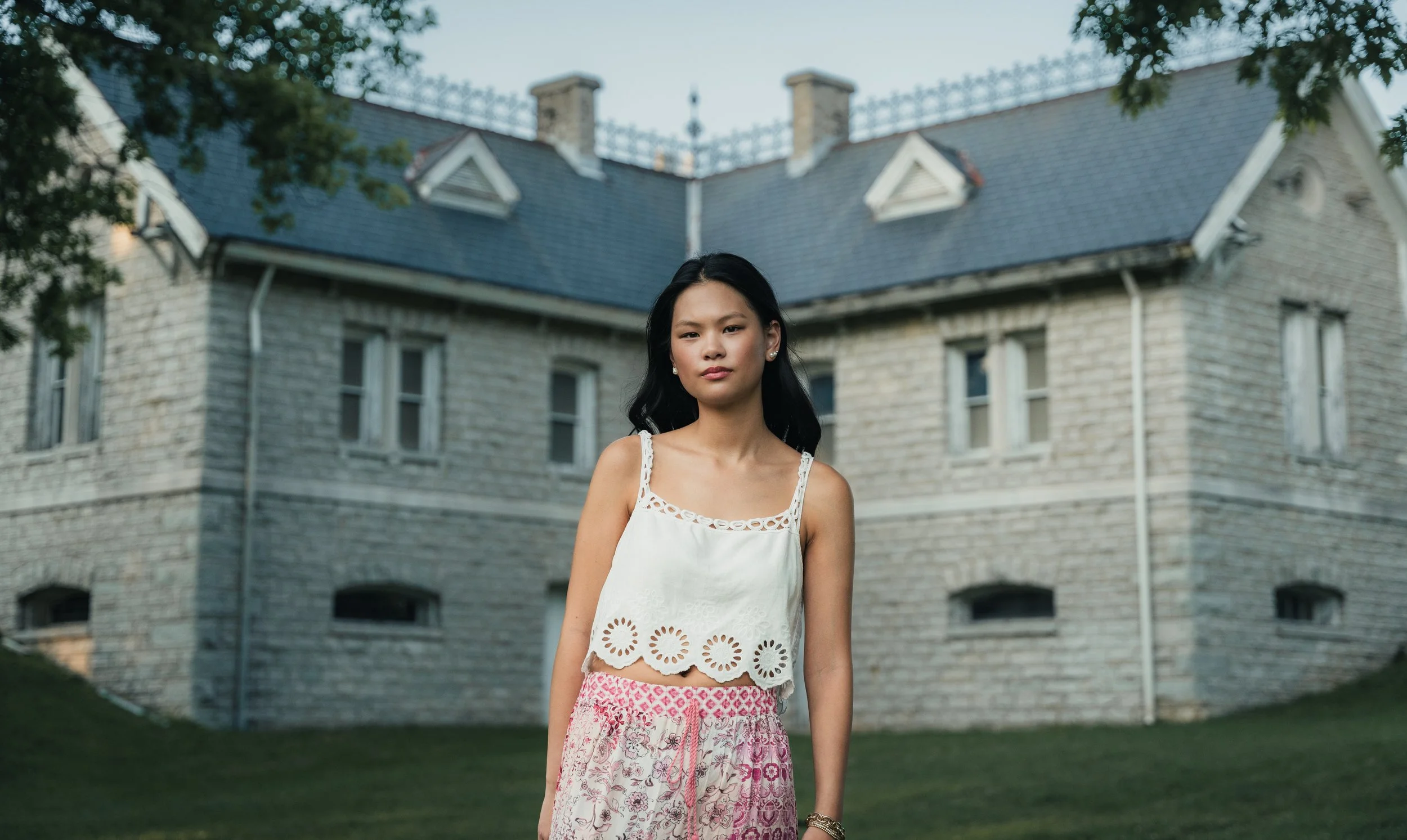 Young woman wearing a white top with eyelet trim and pink patterned skirt standing outdoors in front of a large stone house with a slate roof, surrounded by trees.