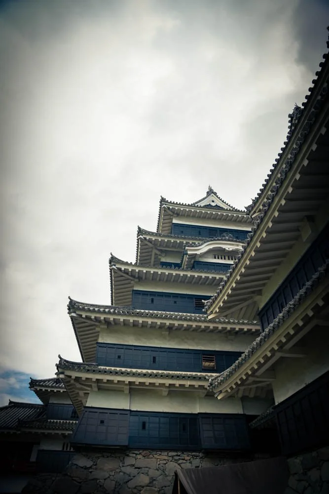 A traditional Japanese castle with multiple tiers and curved rooflines seen from below, against a cloudy sky.