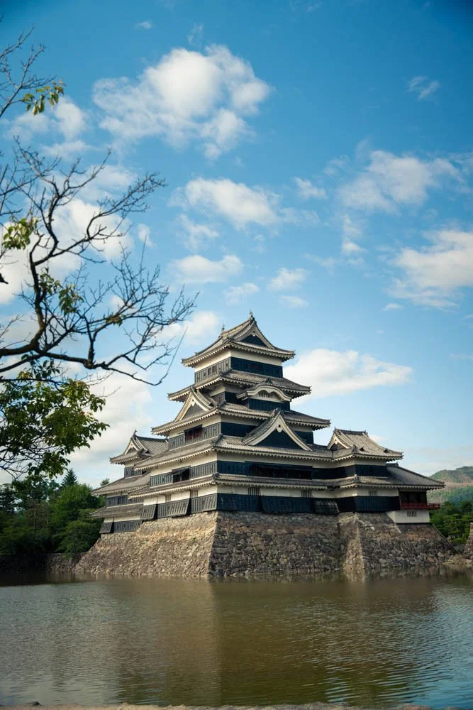 Traditional Japanese castle with multiple tiers surrounded by water and trees, under a blue sky with clouds.