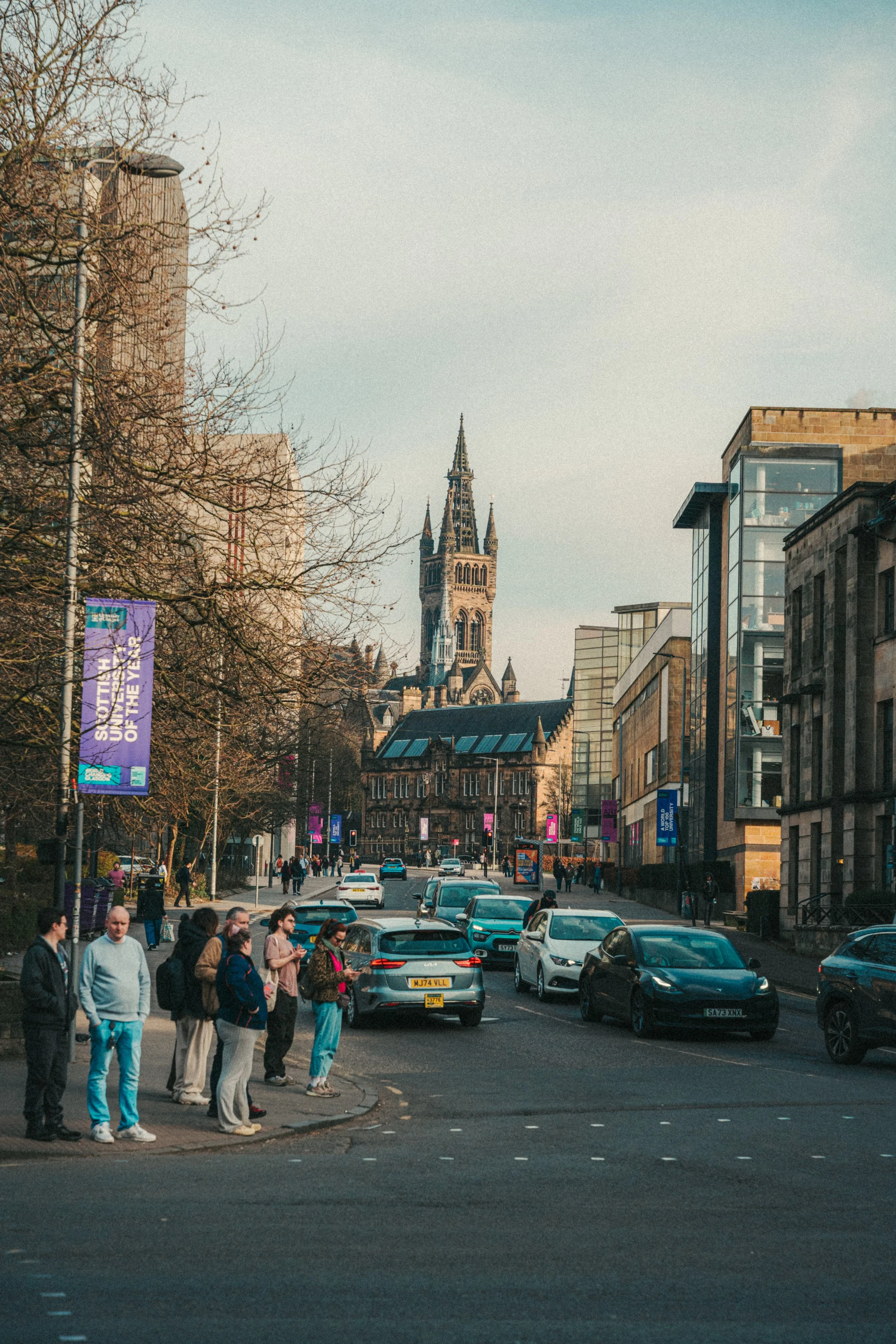 Street scene in a city with cars parked and people crossing the road, with historical and modern buildings, including a tall church tower in the background.