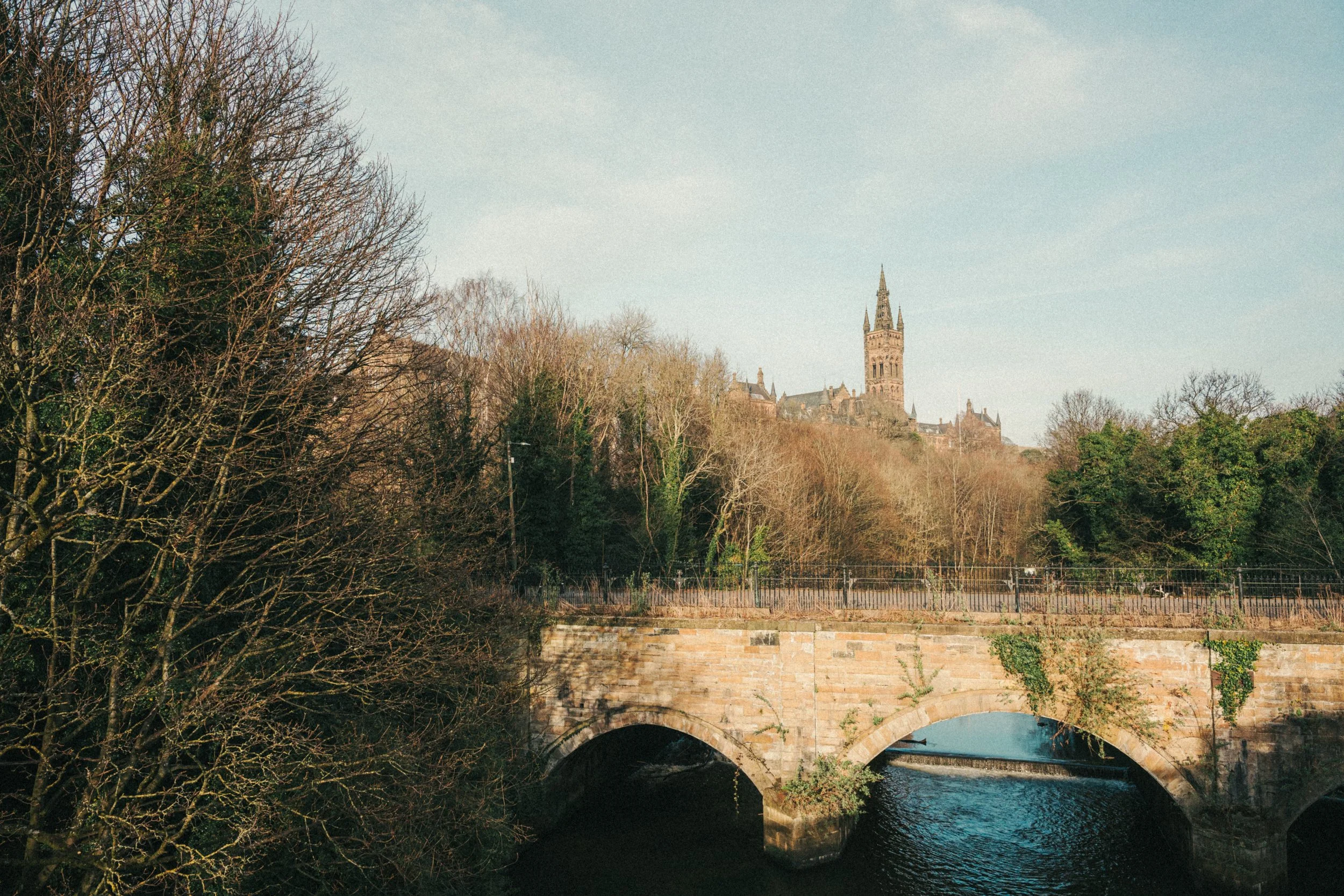 View of historic castle on hilltop with clock tower, surrounded by leafless trees, over a river with stone bridge.
