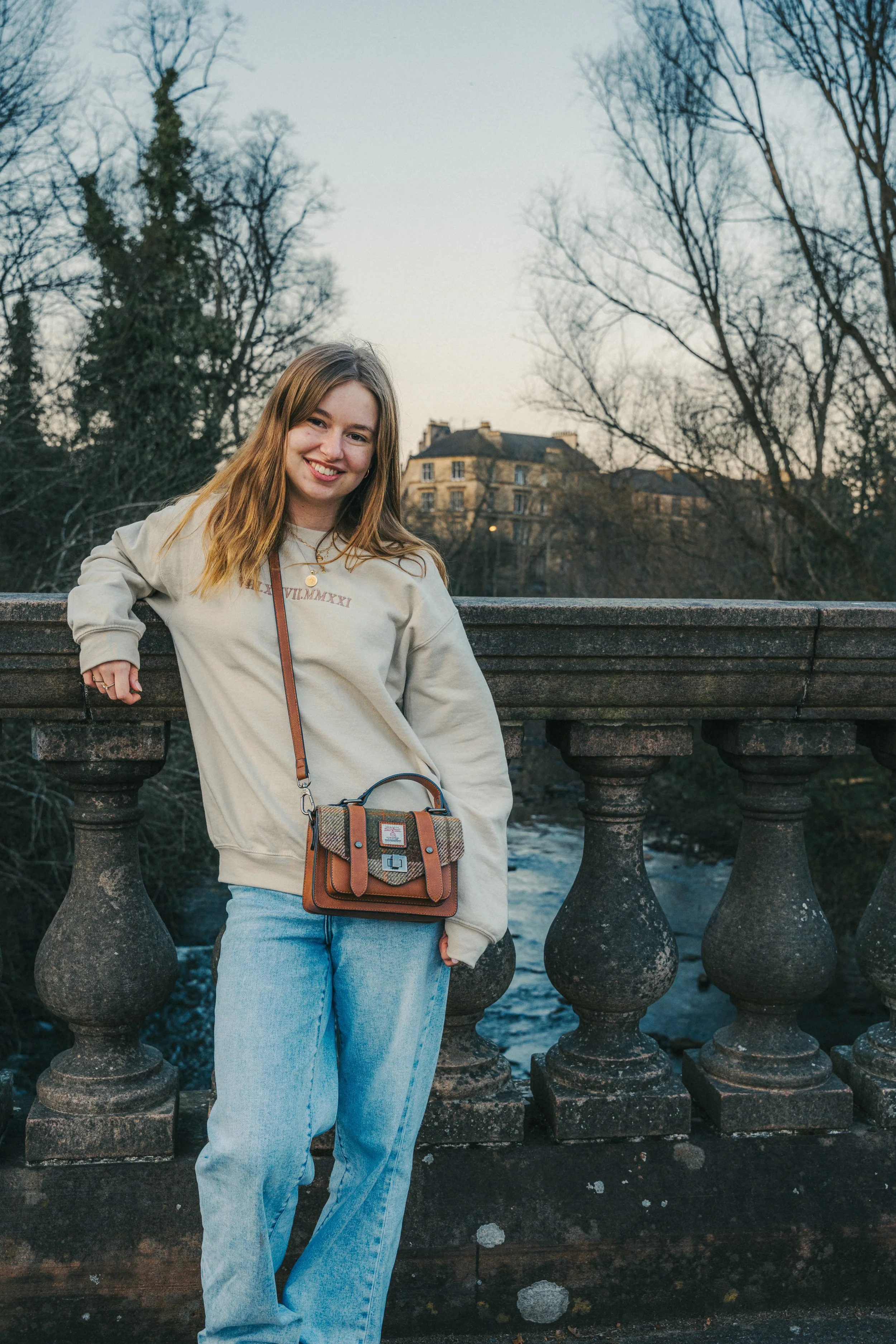 A young woman with long light brown hair smiling, leaning on a stone railing overlooking a river, with leafless trees and a city building in the background during dusk or sunset.