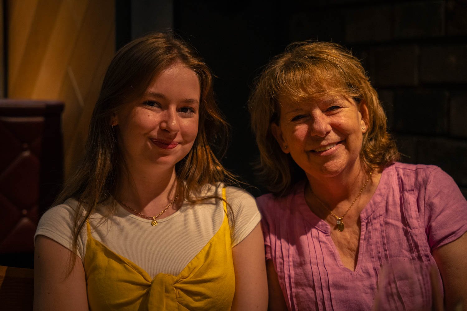 Two women sitting together in a dimly lit setting, smiling at the camera.