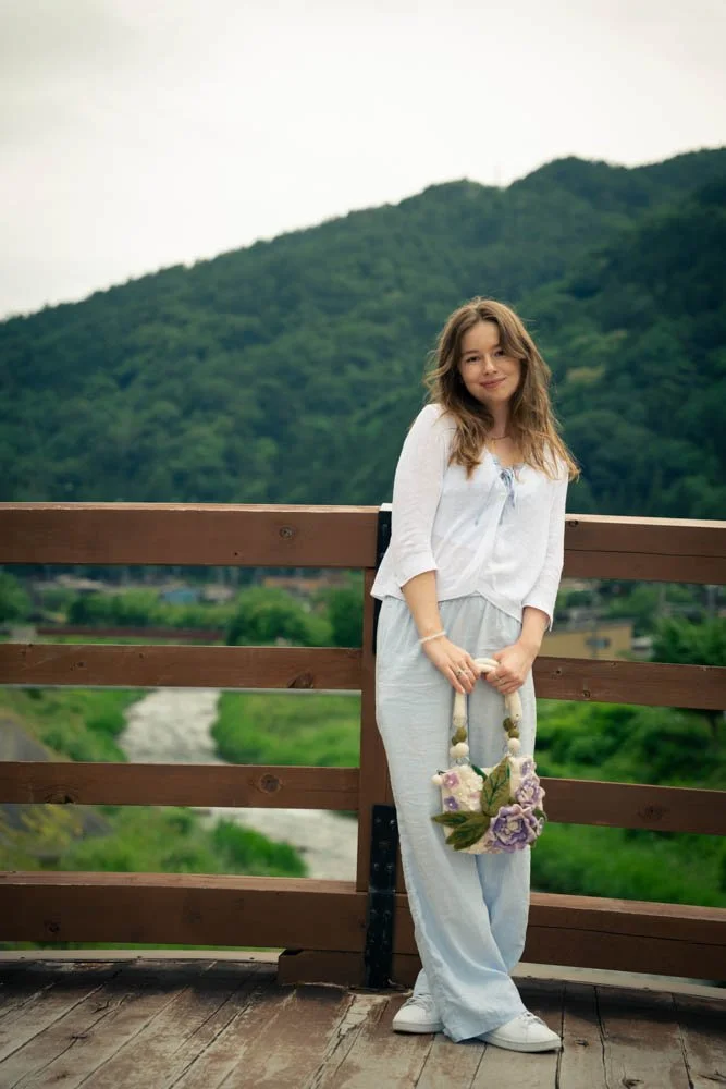 A young woman with long brown hair, dressed in a white top and light gray pants, stands on a wooden bridge holding a floral bouquet, with a green mountain and river in the background.