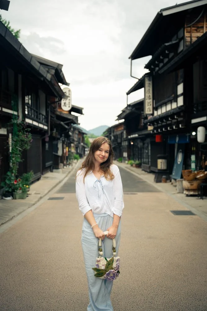 A young woman with light brown hair smiling and holding a floral-colored bag stands in the middle of a quiet traditional Japanese street lined with dark wooden buildings and shops.