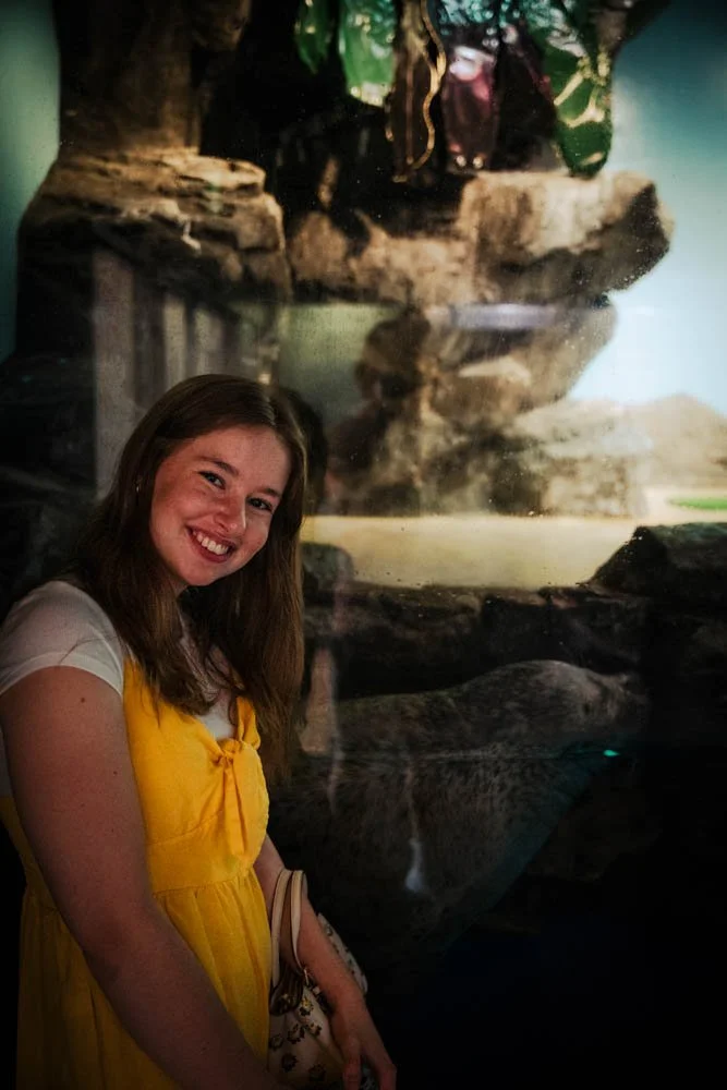 A woman with long brown hair smiling and wearing a yellow dress standing in front of a glass enclosure with a large cat, possibly a snow leopard, inside.