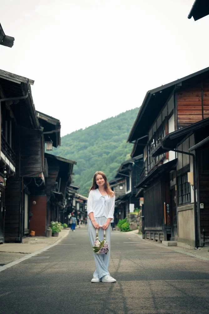 A woman standing in the middle of a quiet street in a traditional Japanese village, holding a bouquet of flowers and smiling, with wooden buildings and a green mountain in the background.