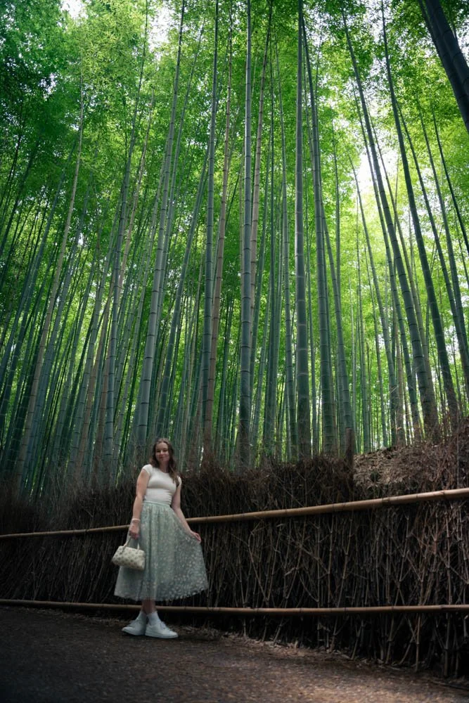 A woman in a white top, light green skirt, and white shoes standing near a bamboo fence in a dense bamboo forest.