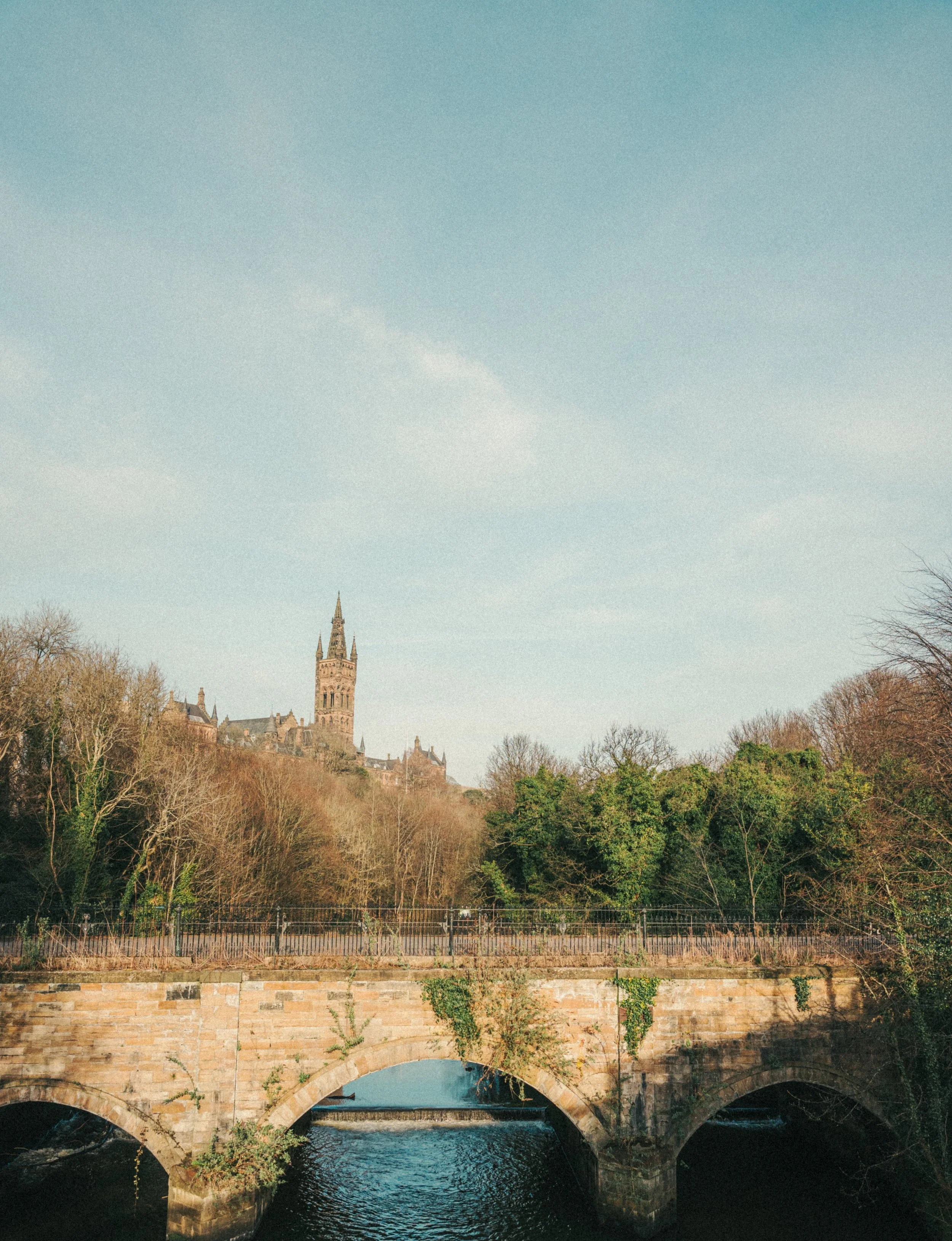 A historic stone bridge with multiple arches over a river, with a towered castle and trees in the background under a blue sky with some clouds.