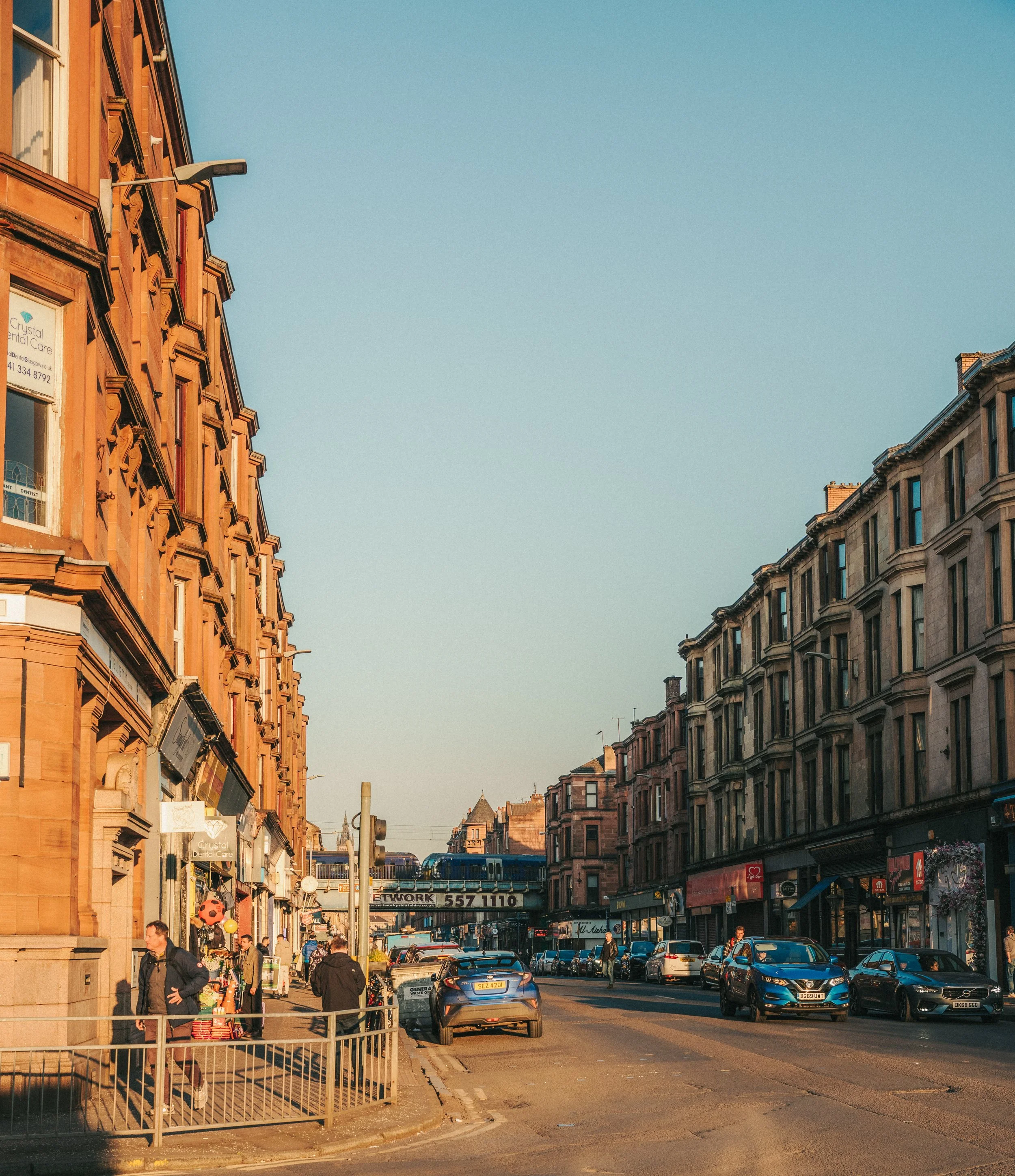 A busy city street lined with old brick buildings, storefronts, and cars parked along the curb. Pedestrians walk on the sidewalk, and there is a street sign across the street. The sky is clear with a soft blue hue.