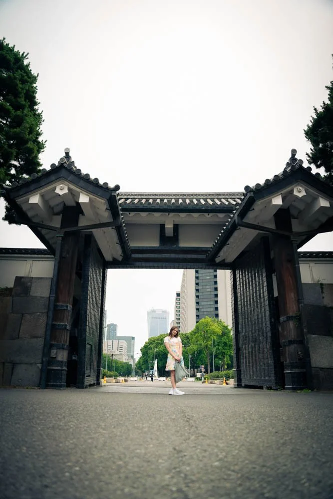 Young woman standing at an open traditional Japanese gate in an urban park with city buildings in the background.