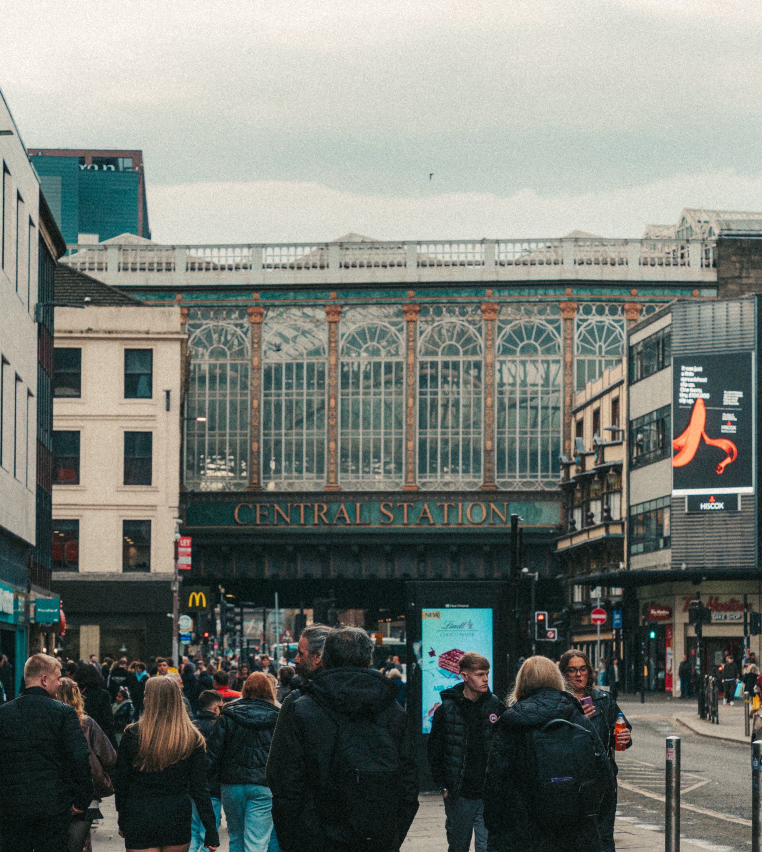 Scene of people walking outdoors near a train station entrance with a sign reading "CENTRAL STATION"; urban area with buildings, billboards, and a crowd of pedestrians.