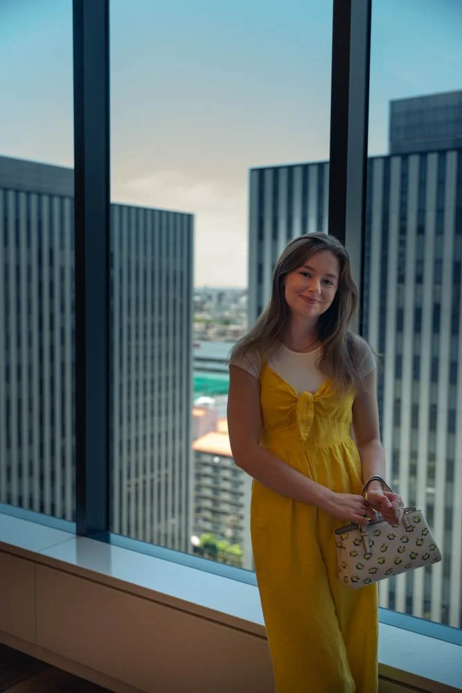 A woman in a yellow dress holding a purse stands by a large window in a high-rise building, with city skyscrapers in the background and partly cloudy sky.