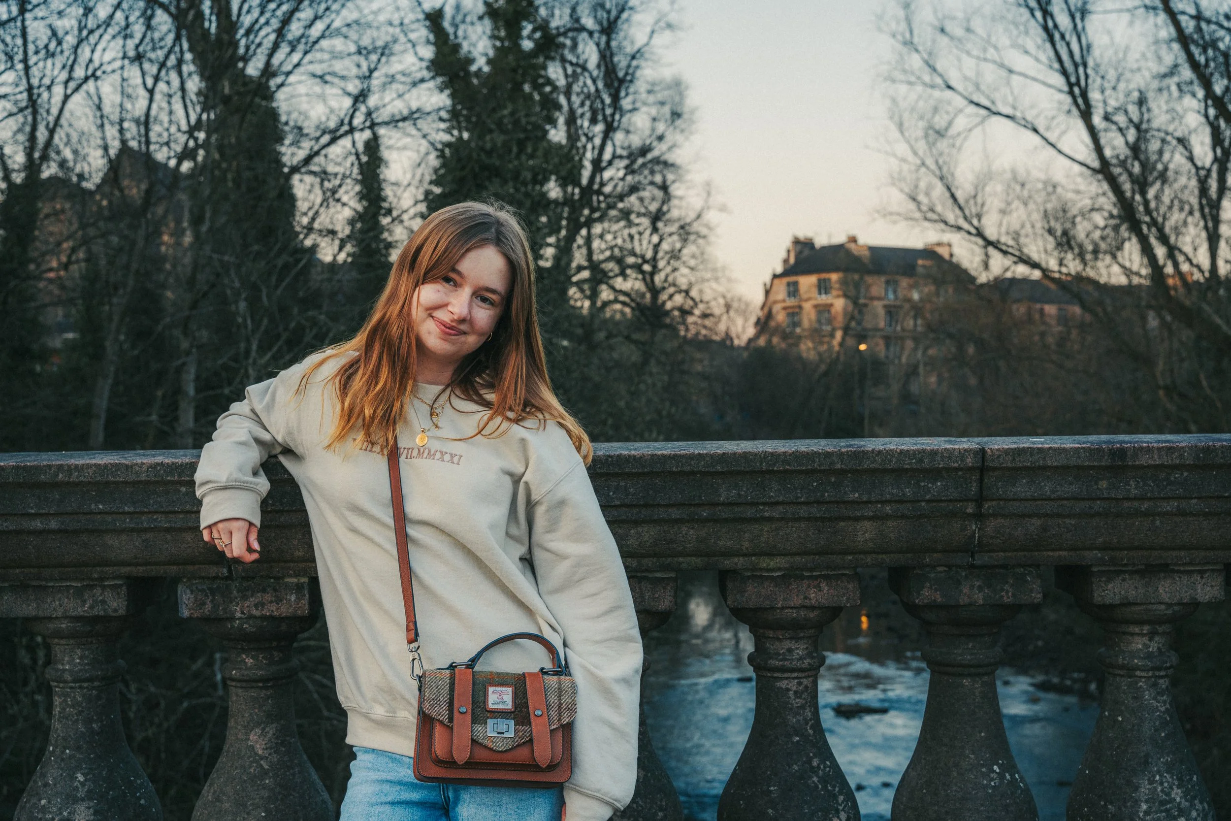 Young woman with light brown hair smiling, standing outdoors by a stone bridge with a river and leafless trees in the background.