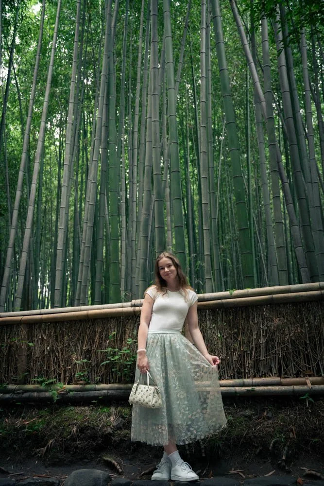 A young woman stands in front of a dense bamboo forest, holding a small handbag and wearing a white top and a sheer, flowy skirt.