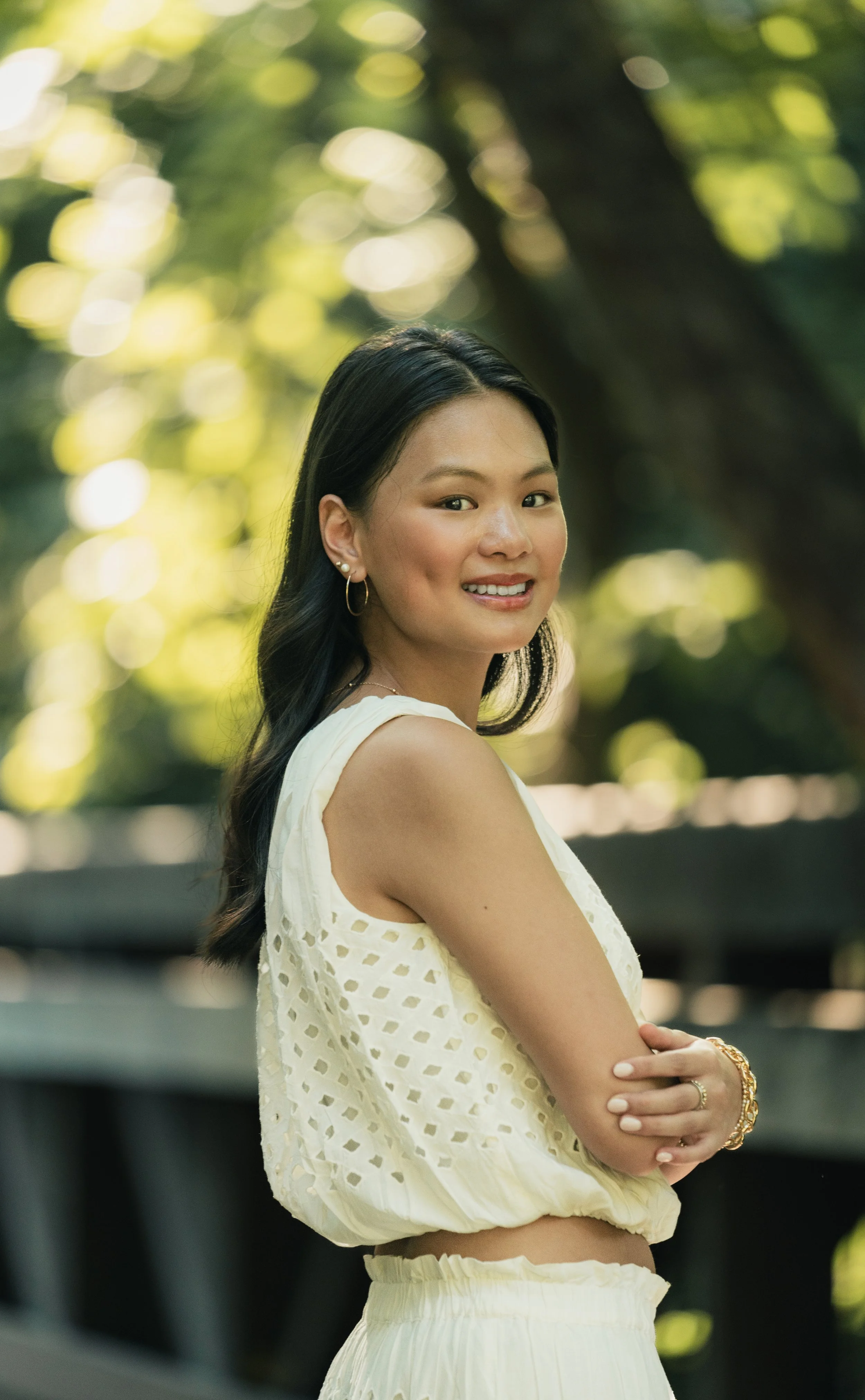 A young woman with long black hair, wearing a white sleeveless top with eyelet details and a matching skirt, is smiling and standing outdoors in a park or garden with blurred green foliage and sunlight in the background.