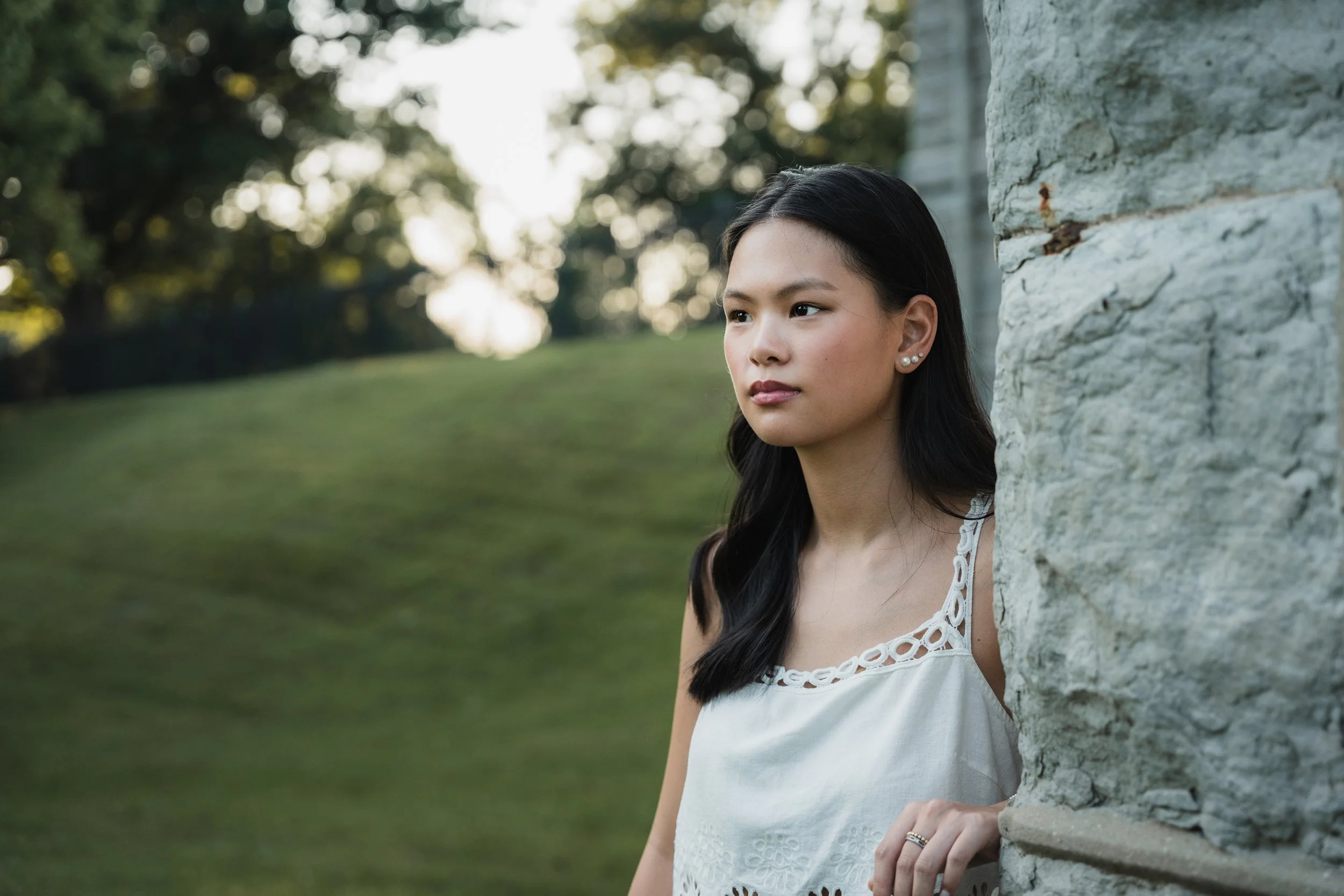 Young woman with long black hair, wearing pearl earrings and a white dress, standing outdoors and leaning against a stone structure.
