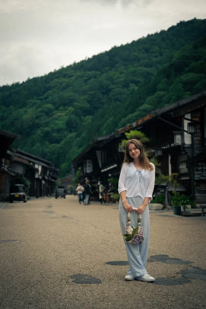 A woman standing on a quiet street in a mountain town, holding a flower basket, with traditional wooden buildings and green mountains in the background, and a few people walking in the distance.