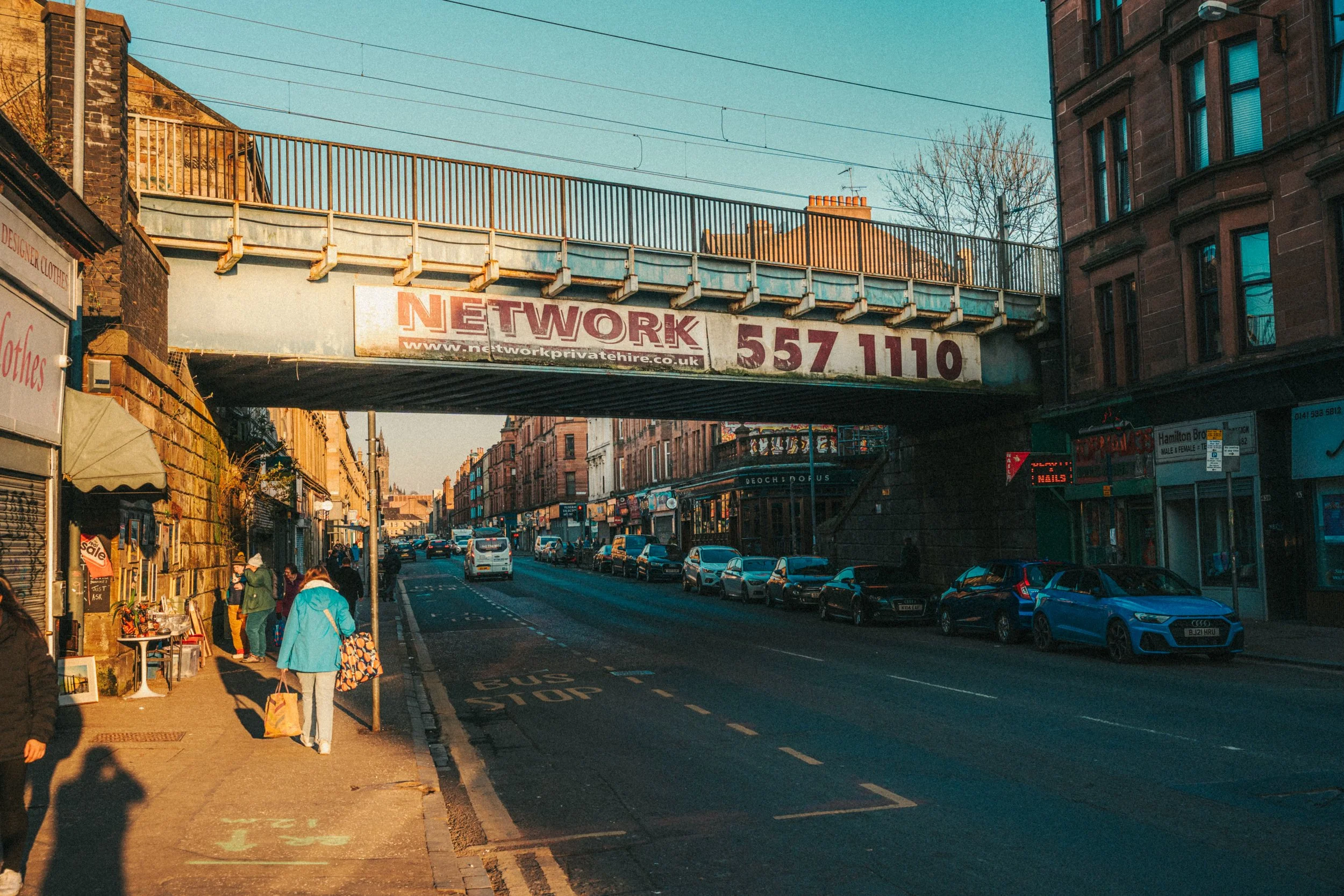 Urban street scene during daytime with cars parked along the street, pedestrians walking on the sidewalk, storefronts, and an elevated train track with a sign displaying a website and phone number.
