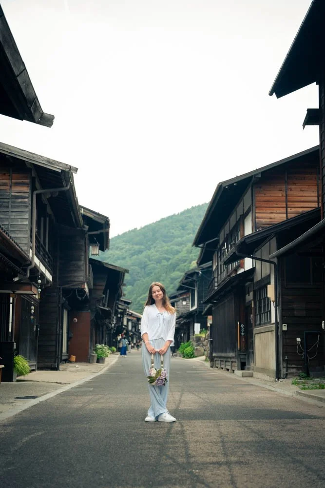 A woman stands in the middle of a deserted street in a traditional Japanese village, holding a bouquet of flowers, with wooden houses on both sides and a mountain in the background.