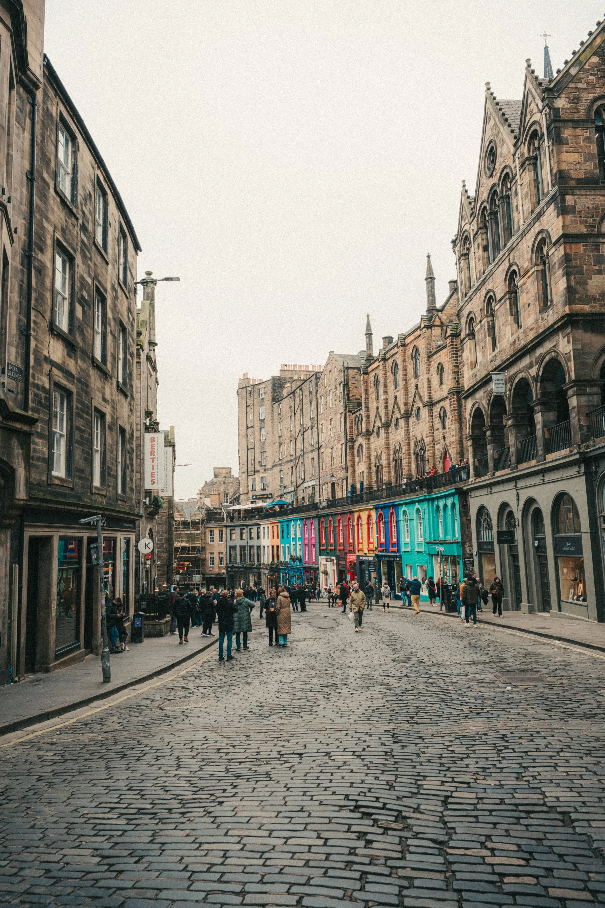 A cobblestone street in a historic city with multi-story stone buildings, some with rounded arches and towers, and people walking. Brightly colored storefronts line the street with a crowd of shoppers and tourists.