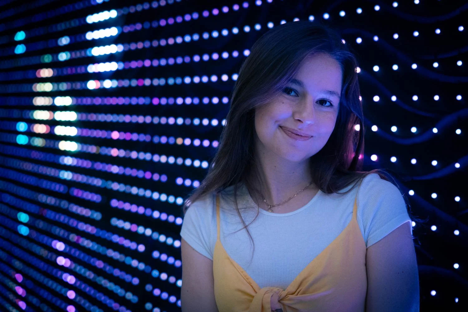 A young woman with long brown hair smiling in front of a background of colorful, glowing LED lights.