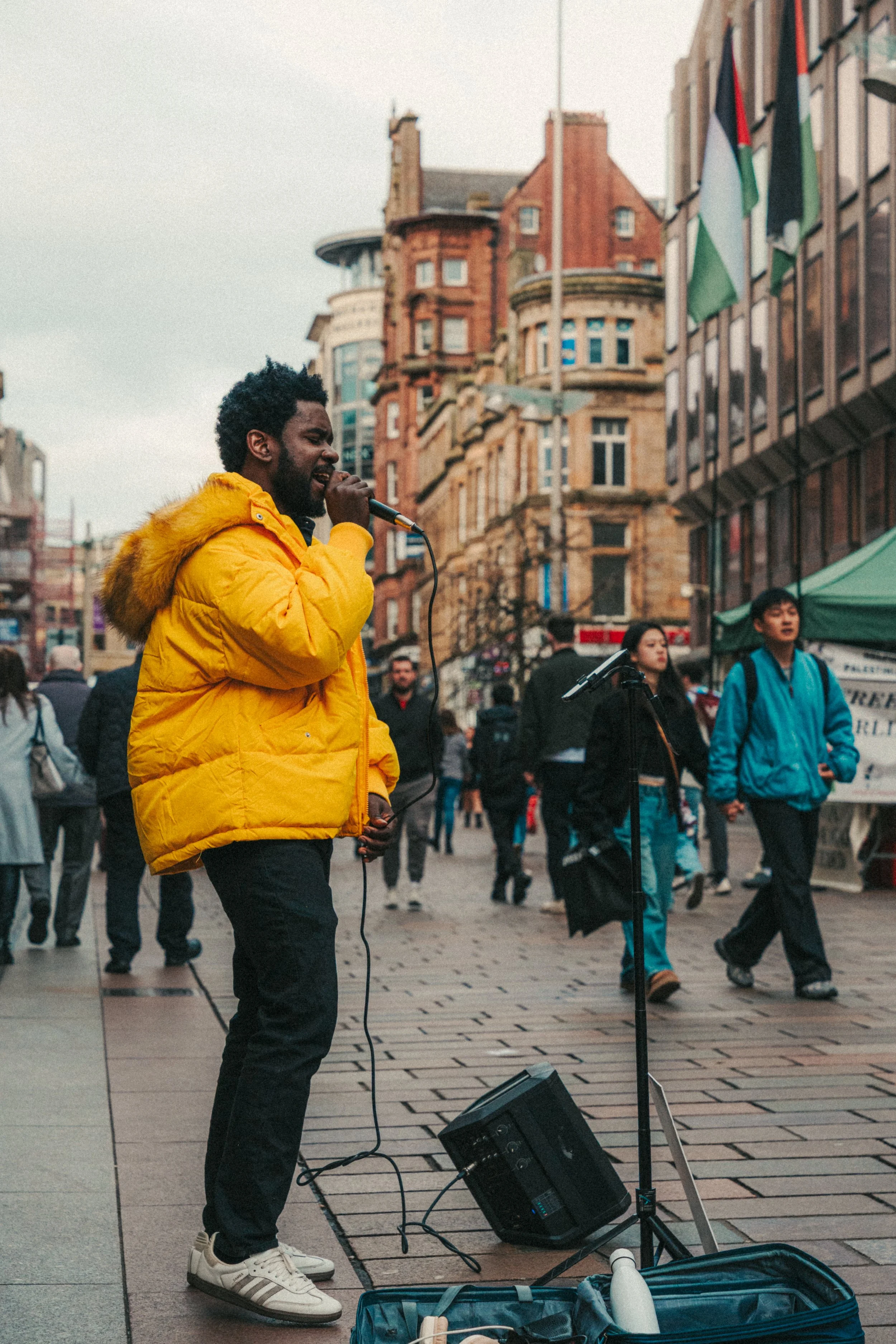 A man in a yellow jacket singing into a microphone on a city street with pedestrians and historic buildings.