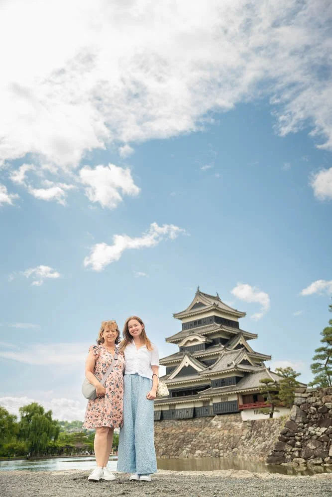 Two women, one older and one younger, standing outdoors in front of a traditional Japanese castle with a stone wall and a body of water, under a partly cloudy sky.