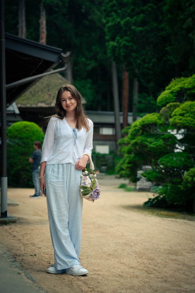 A young woman with long hair wearing a white top and white pants, standing outdoors in a garden with green trees and bushes, holding a bouquet of flowers.