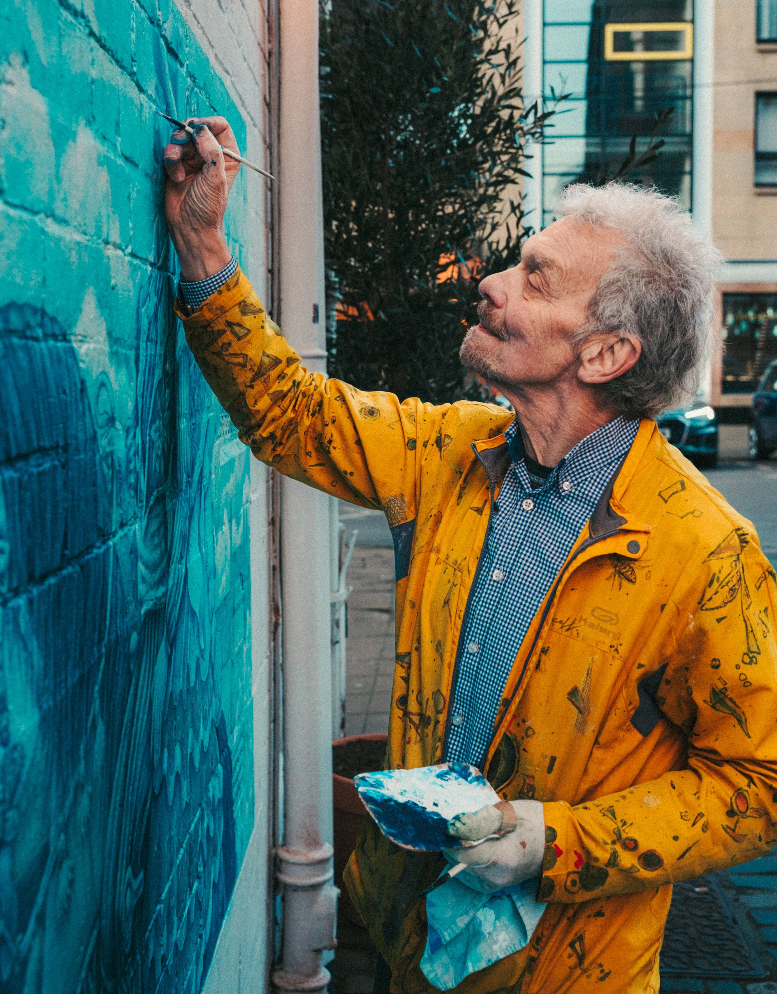 An elderly man with curly gray hair and a beard painting a mural on a brick wall with a paintbrush, wearing a yellow painter's jacket with paint splatters and a checkered shirt underneath, holding a palette in his other hand.