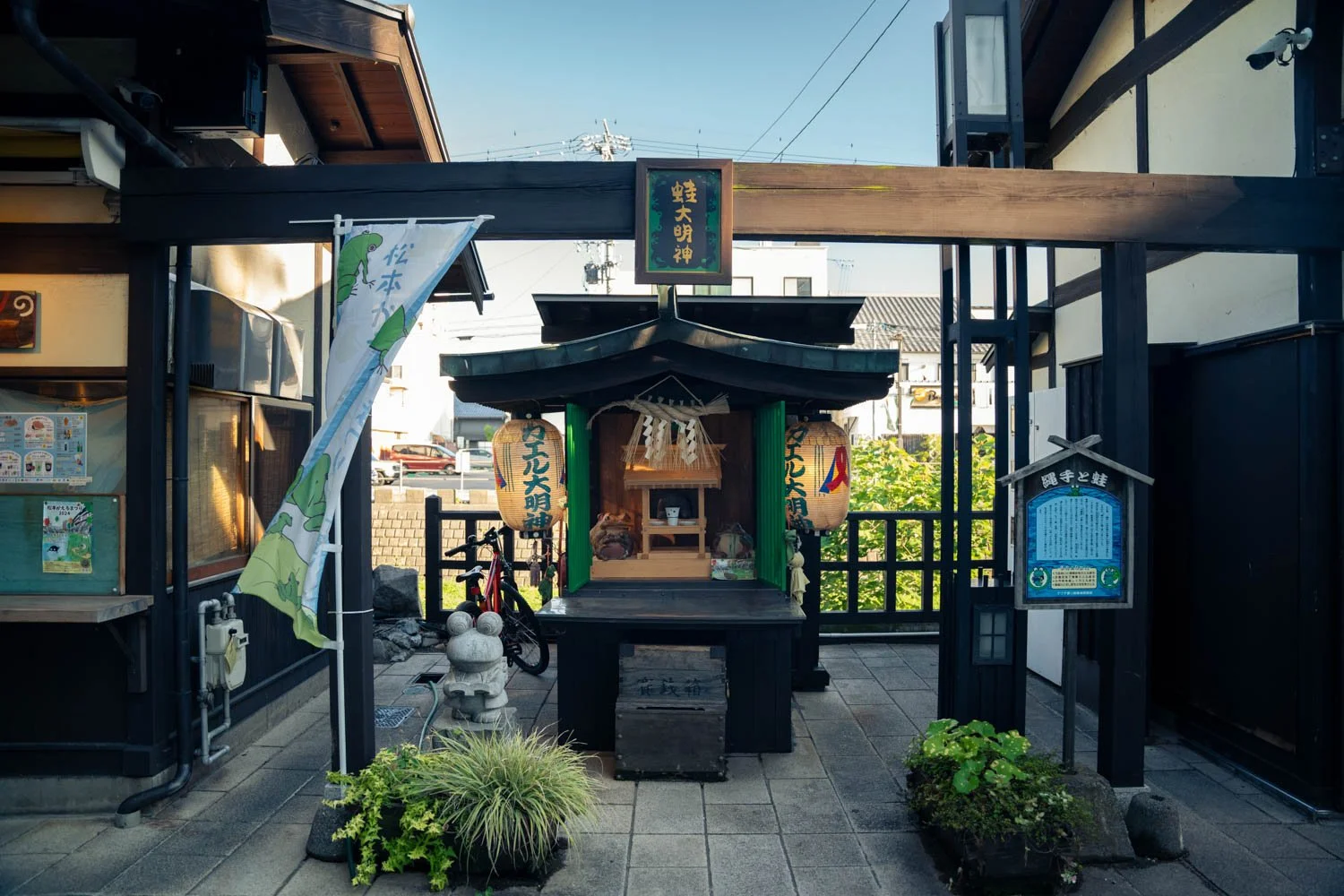 A small Japanese shrine with lanterns, wooden structure, and offerings, situated between two buildings on a paved area, with plants in front and a bicycle in the background.