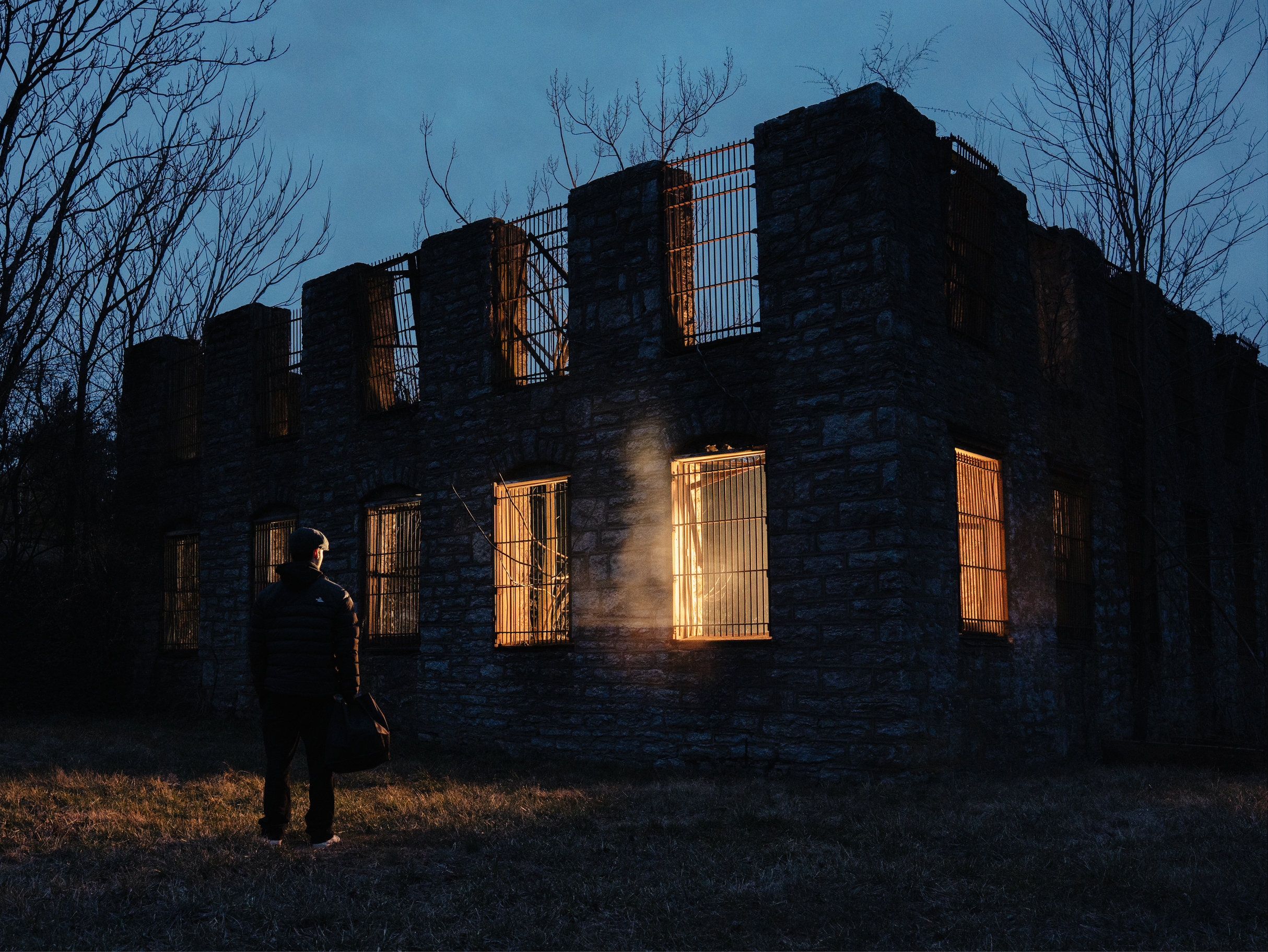 A person holds a long stick and stands in front of a burning building made of stone, with fire visible through the windows, during twilight or early night, with bare trees surrounding the structure.