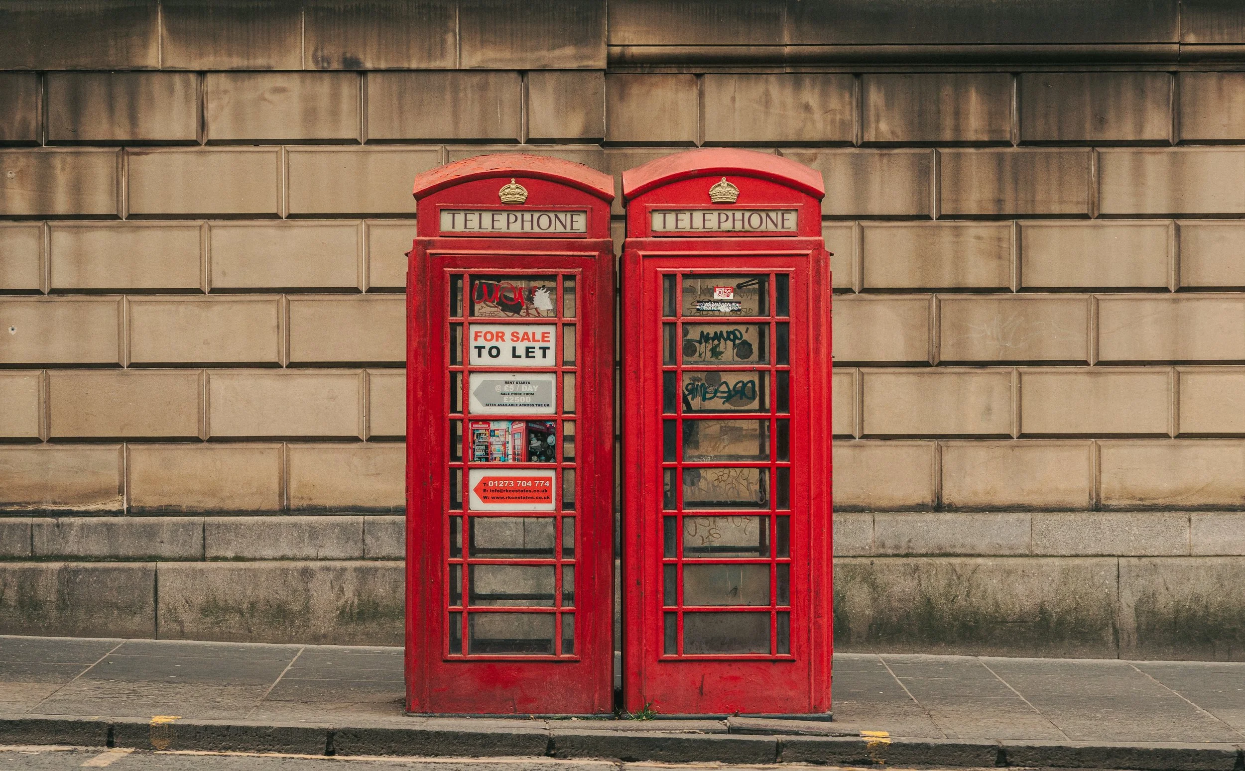 Two traditional red British telephone booths standing side by side against a beige brick wall, with graffiti on the glass panes, and a black sidewalk and curb in the foreground.