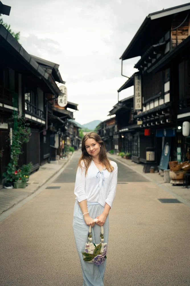 A woman standing in the middle of a traditional Japanese street, holding a bag decorated with flowers, with wooden buildings and shop signs on both sides.